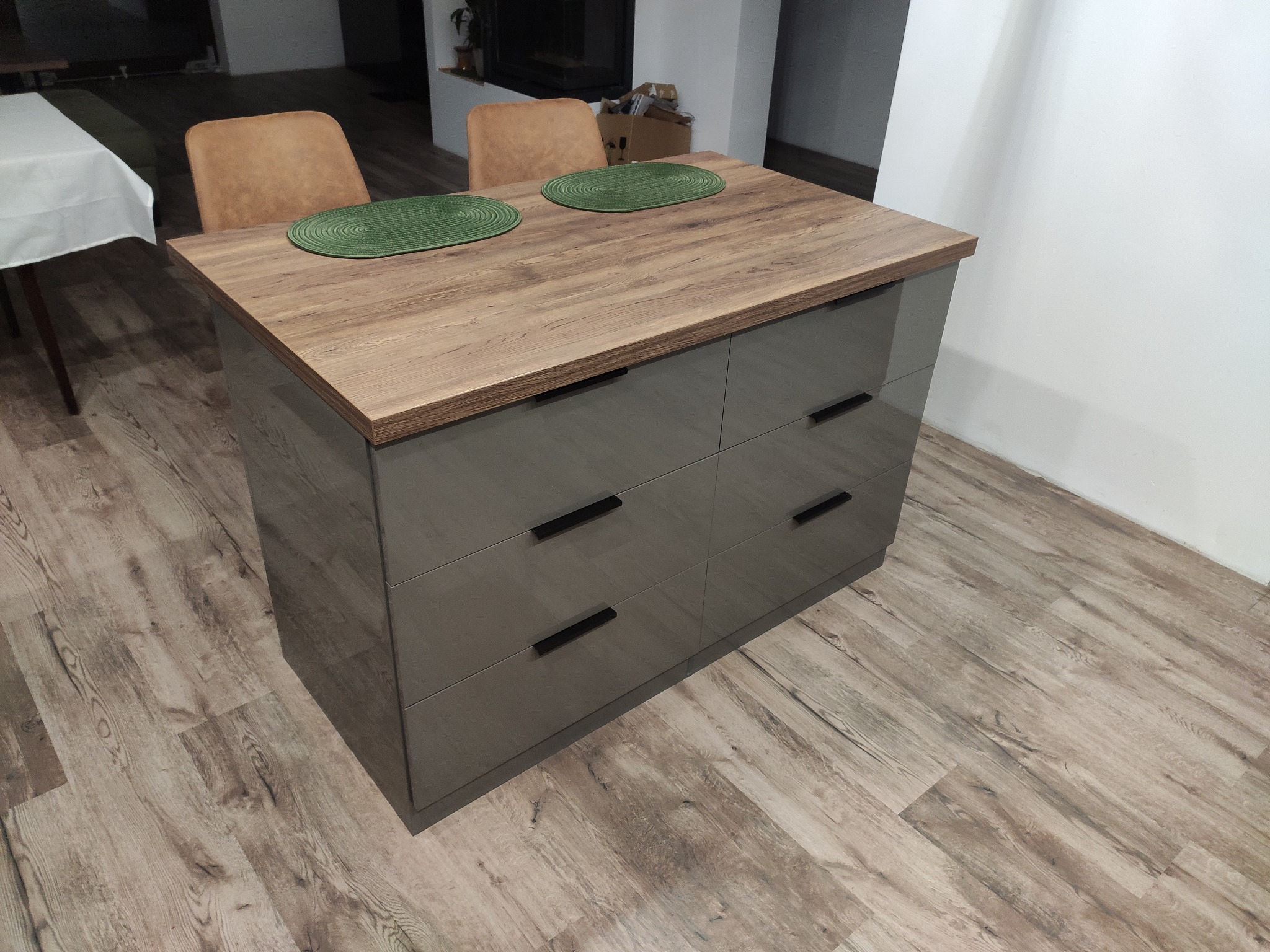 Wyspa kuchenna z blatem imitującym drewno i szarymi, lakierowanymi frontami szuflad, uzupełniona o dwa okrągłe, zielone podkładki na blacie, stojąca na podłodze z paneli imitujących drewno.