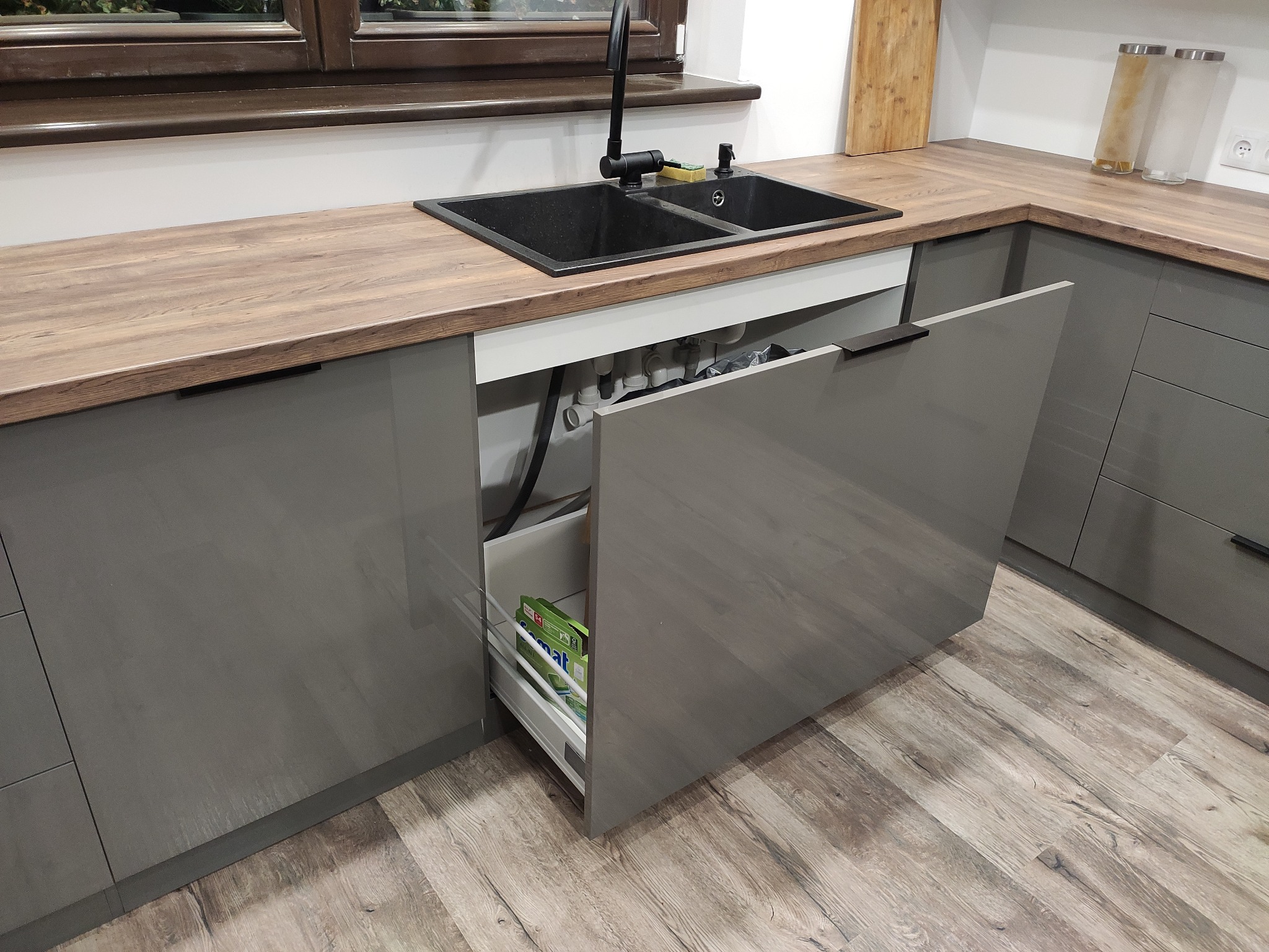 Szara kuchnia z drewnianym blatem, czarnym zlewem i baterią, z otwartą szafką pod zlewem, w której widoczny jest syfon i środki czystości.