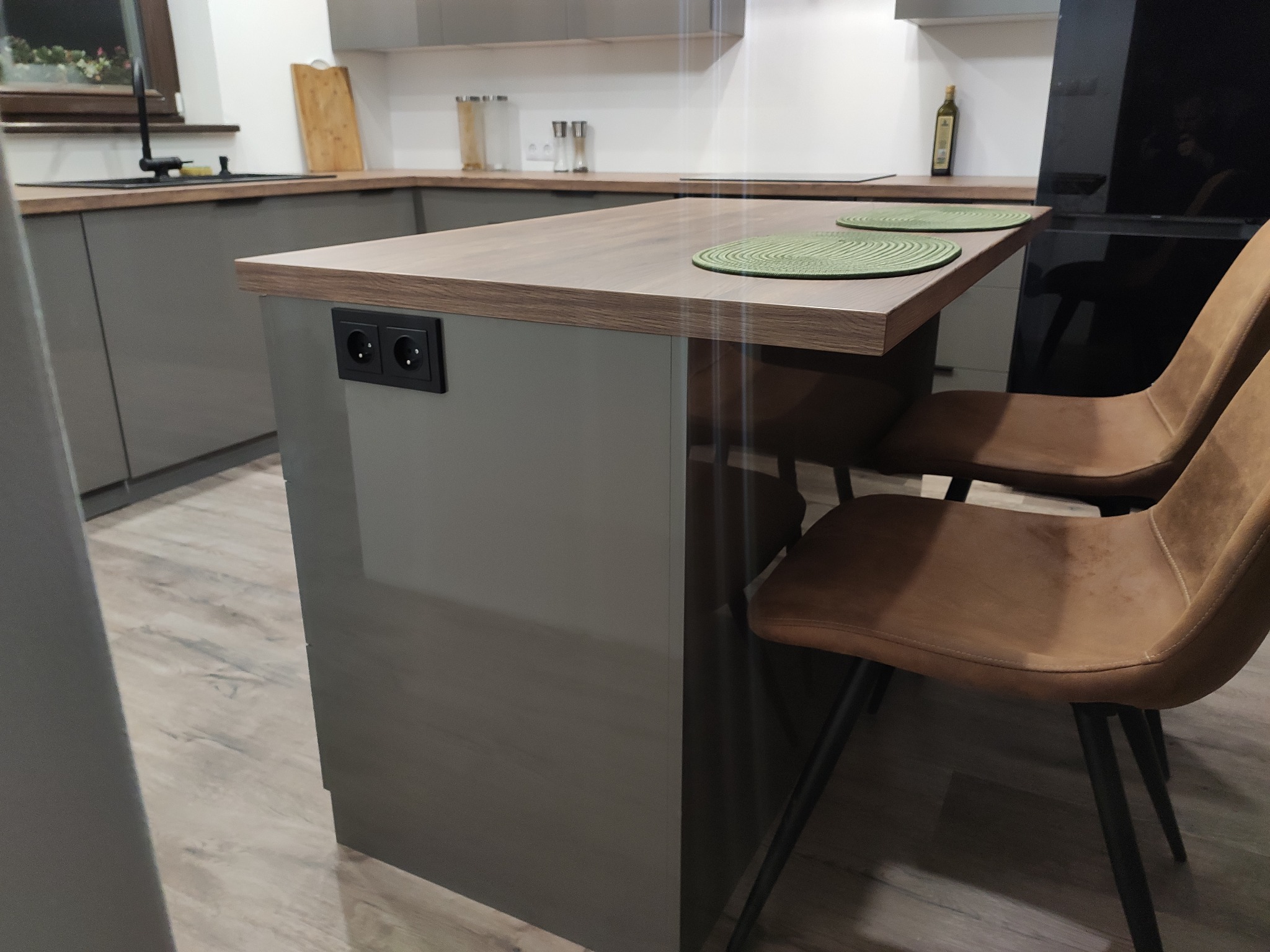 Szara kuchnia z wyspą i drewnianym blatem, widoczne dwa brązowe krzesła, gniazdka elektryczne w wyspie, oraz zielone podkładki na blacie.