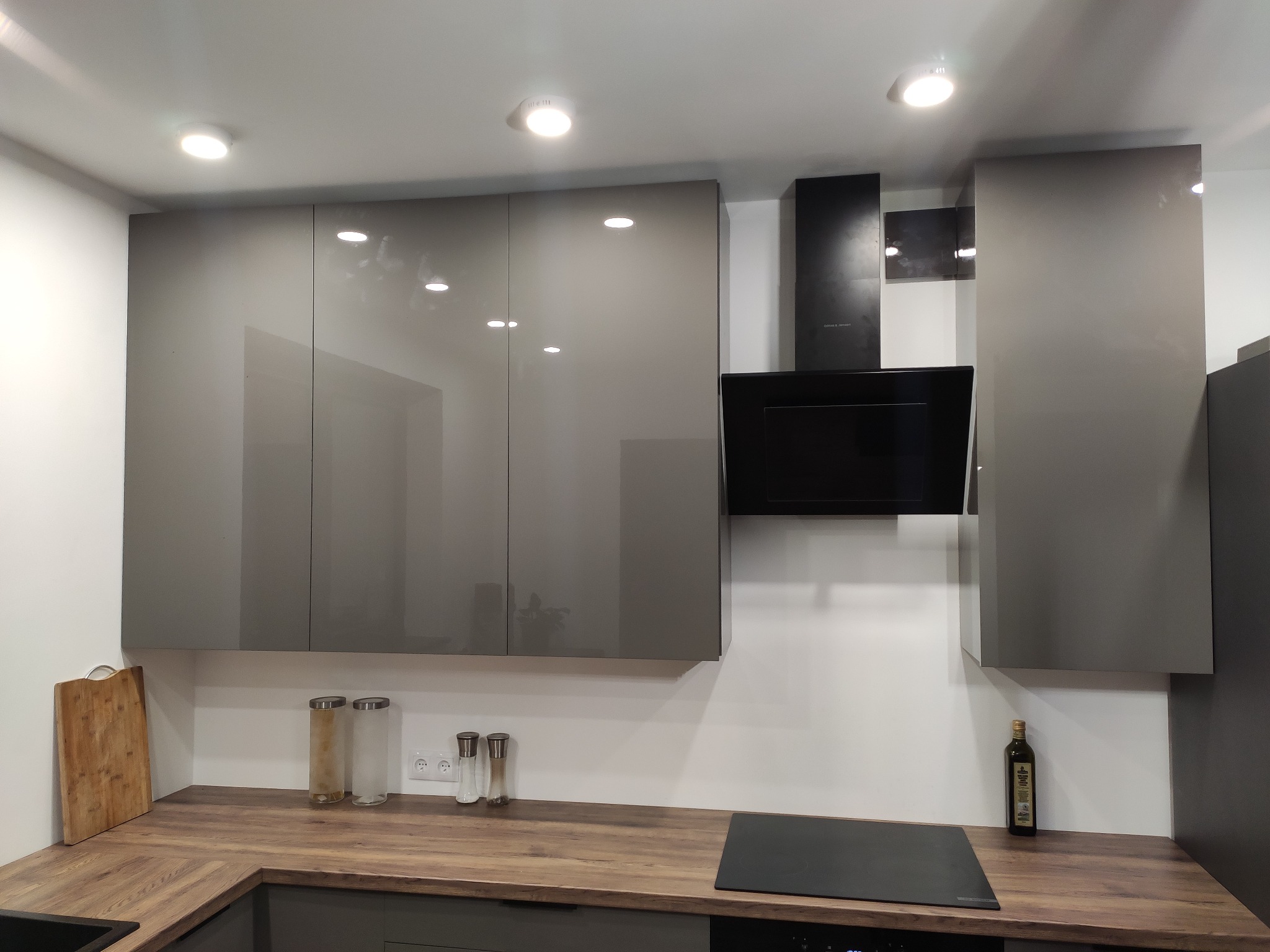 Nowoczesna kuchnia: szare, lakierowane szafki wiszące, drewnopodobny blat, czarny okap i płyta indukcyjna, minimalistyczne dodatki.