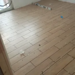 Układanie podłogi z płytek imitujących drewno w jasnym odcieniu, z użyciem poziomicy i krzyżyków dystansowych, widoczne niedokończone fragmenty przy progu.