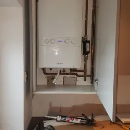 Biały piec gazowy marki Ideal Logic+ zamontowany w szafce kuchennej, widoczne miedziane rury i narzędzia hydrauliczne na blacie, w tym wiertarka akumulatorowa i pistolet do silikonu.