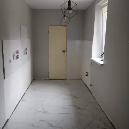 Wnętrze w trakcie remontu: położona marmurowa podłoga, częściowo wykończone ściany z fragmentami płytek, surowy stan ścian w szarym odcieniu, geometryczna lampa wisząca, widoczne instalacje pod oknem.