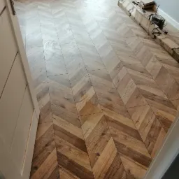 Ułożone panele podłogowe w jodełkę, widoczne opakowania po panelach, fragment drzwi i okna.