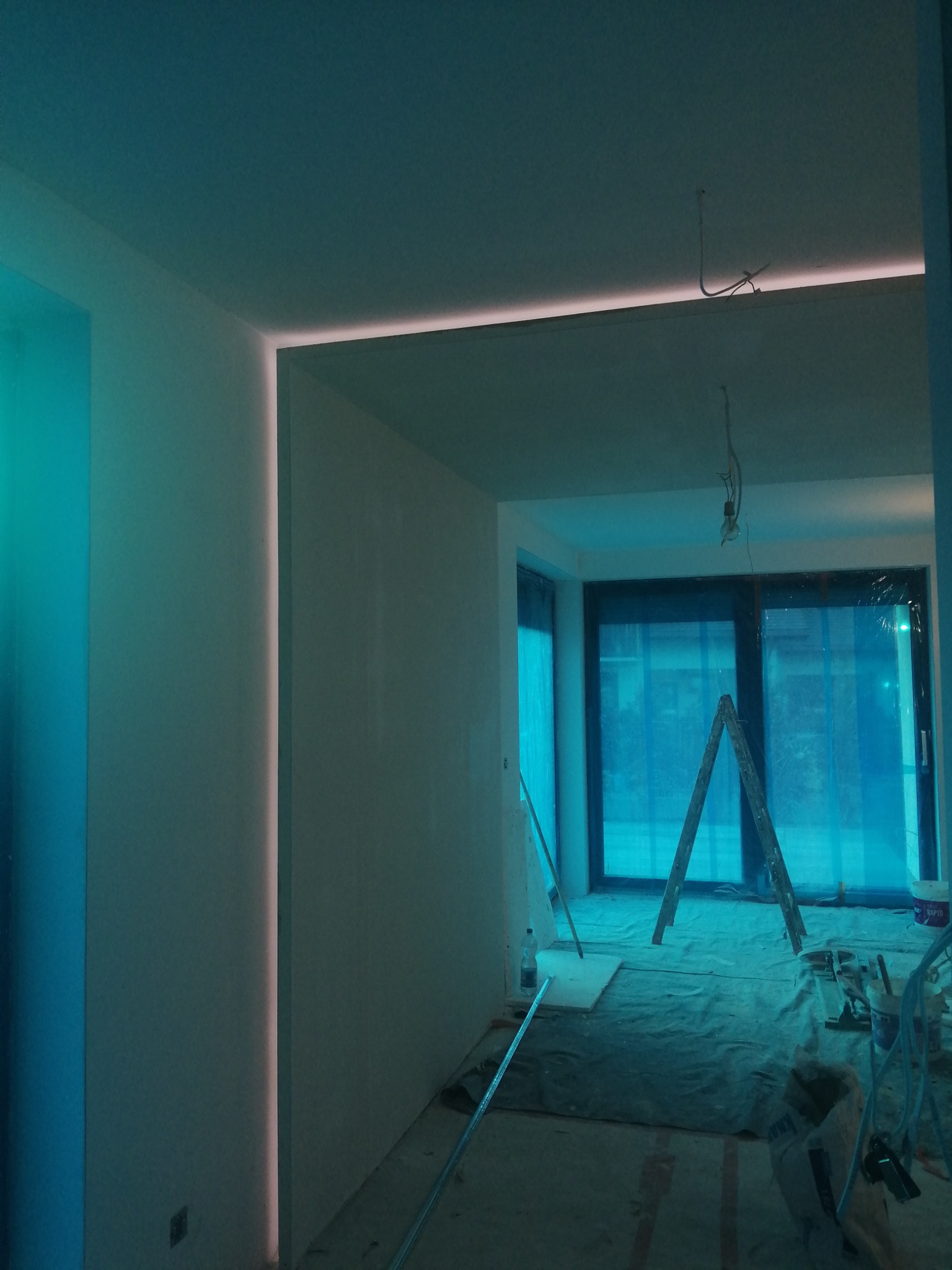 Wnętrze w trakcie prac wykończeniowych, widoczne ściany przygotowane pod malowanie, listwy oświetleniowe LED w suficie i ścianach, zabezpieczona folią podłoga i okna, drabina oparta o okno.