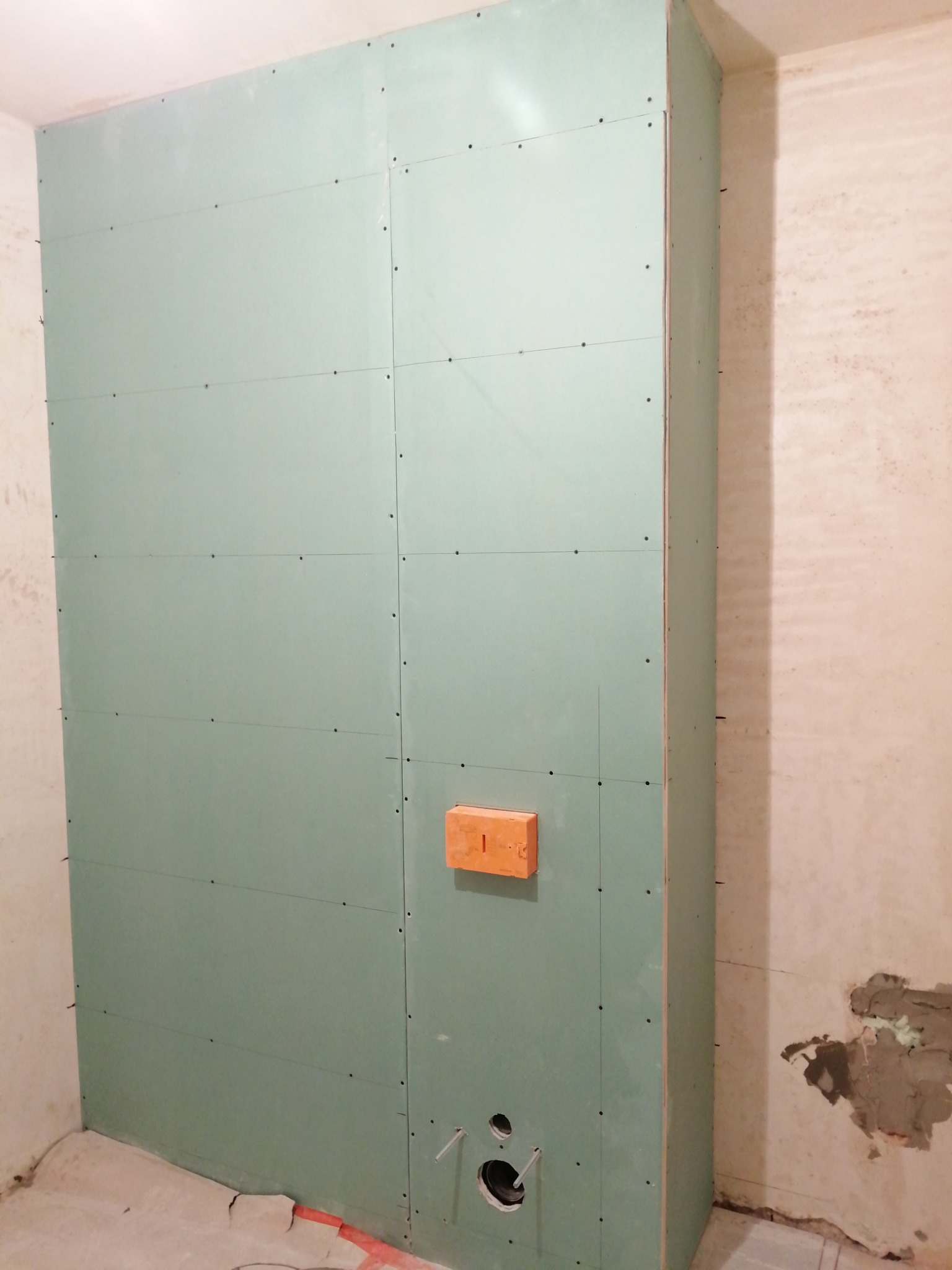 Ściana z zielonej płyty gipsowo-kartonowej zamontowana w rogu pomieszczenia, widoczne otwory na instalacje i pomarańczowa puszka elektryczna, ślady po zaprawie na ścianie i podłodze.