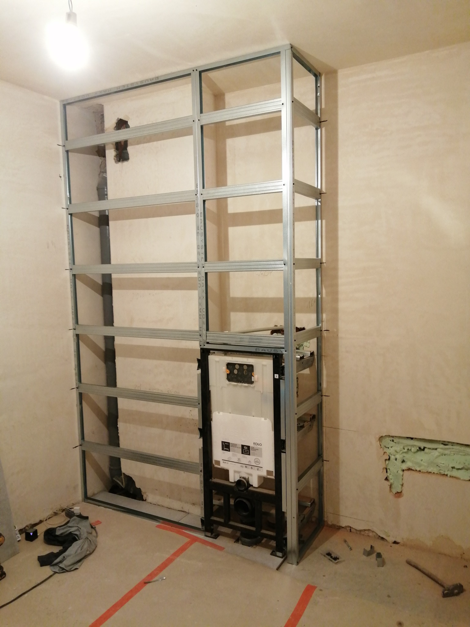 Stelaż z profili metalowych do zabudowy podtynkowego WC, widoczne rury kanalizacyjne i instalacja wodna, ściany w stanie surowym, podłoga z wyznaczonymi liniami, etap prac remontowych w łazience.