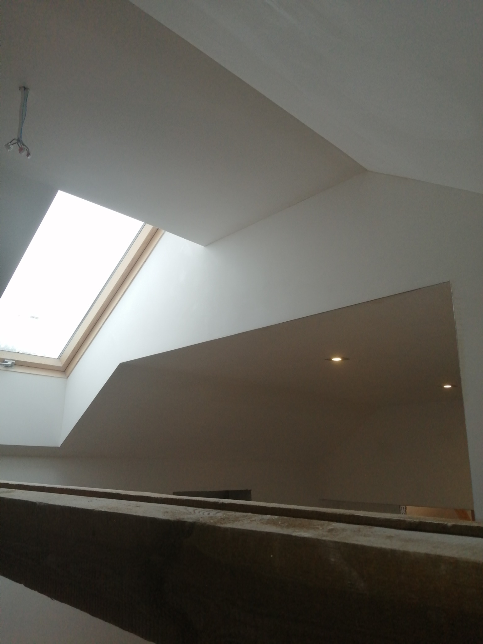 Gładkie, białe ściany i sufit poddasza z oknem dachowym i wpuszczanymi lampami, widoczne z dołu przez drewnianą konstrukcję.