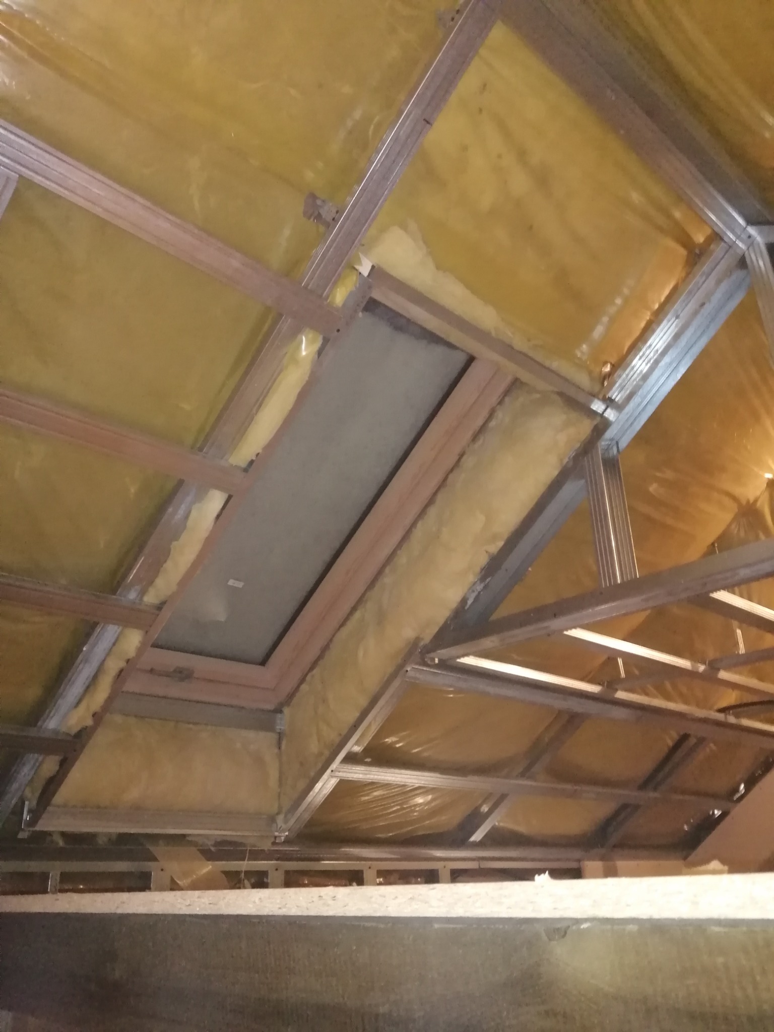 Szkielet sufitu podwieszanego z profilami metalowymi i izolacją z wełny mineralnej, widoczne fragmenty płyty kartonowo-gipsowej w otworze.