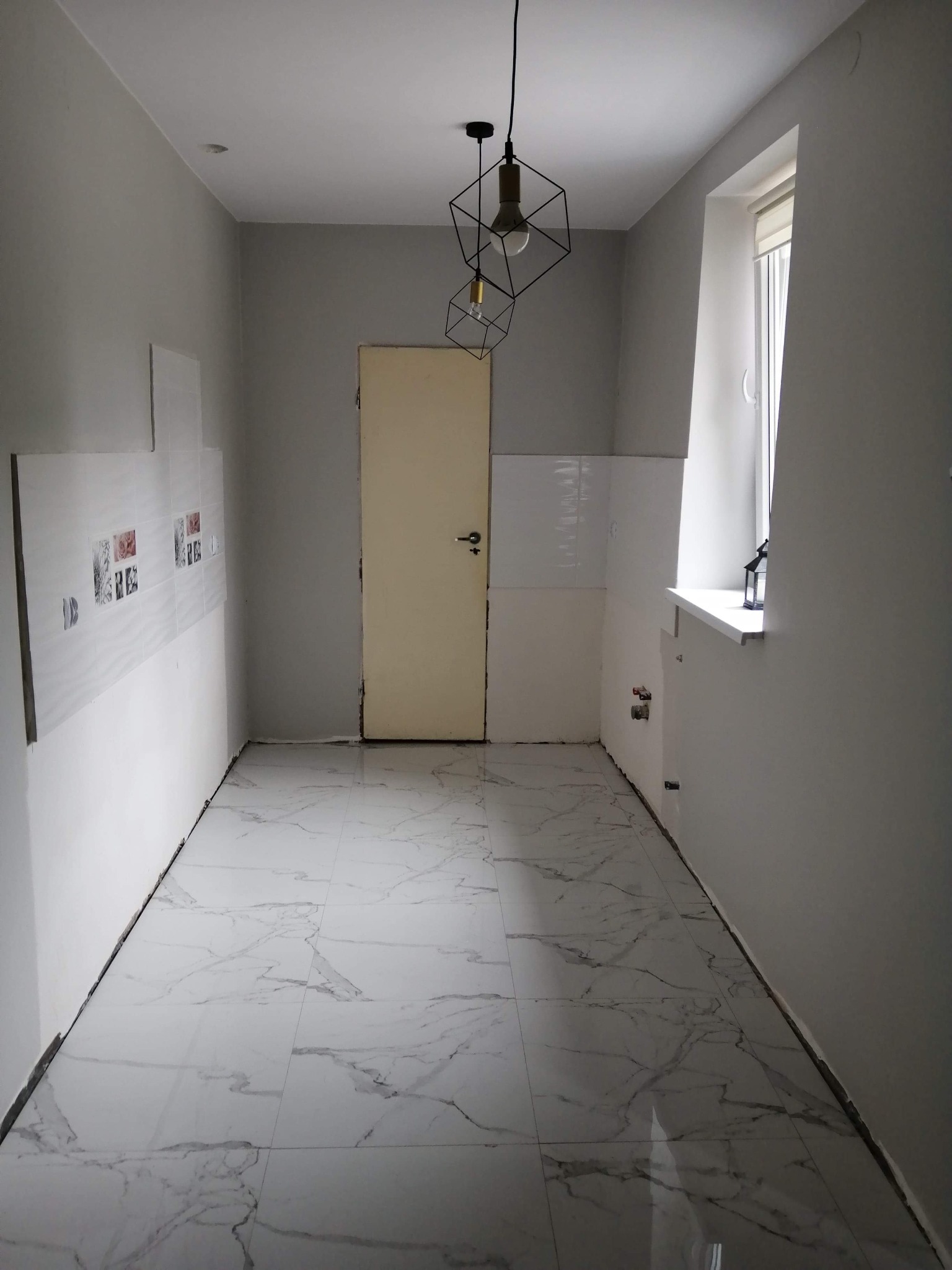 Wnętrze w trakcie remontu: położona marmurowa podłoga, częściowo wykończone ściany z fragmentami płytek, surowy stan ścian w szarym odcieniu, geometryczna lampa wisząca, widoczne instalacje pod oknem.
