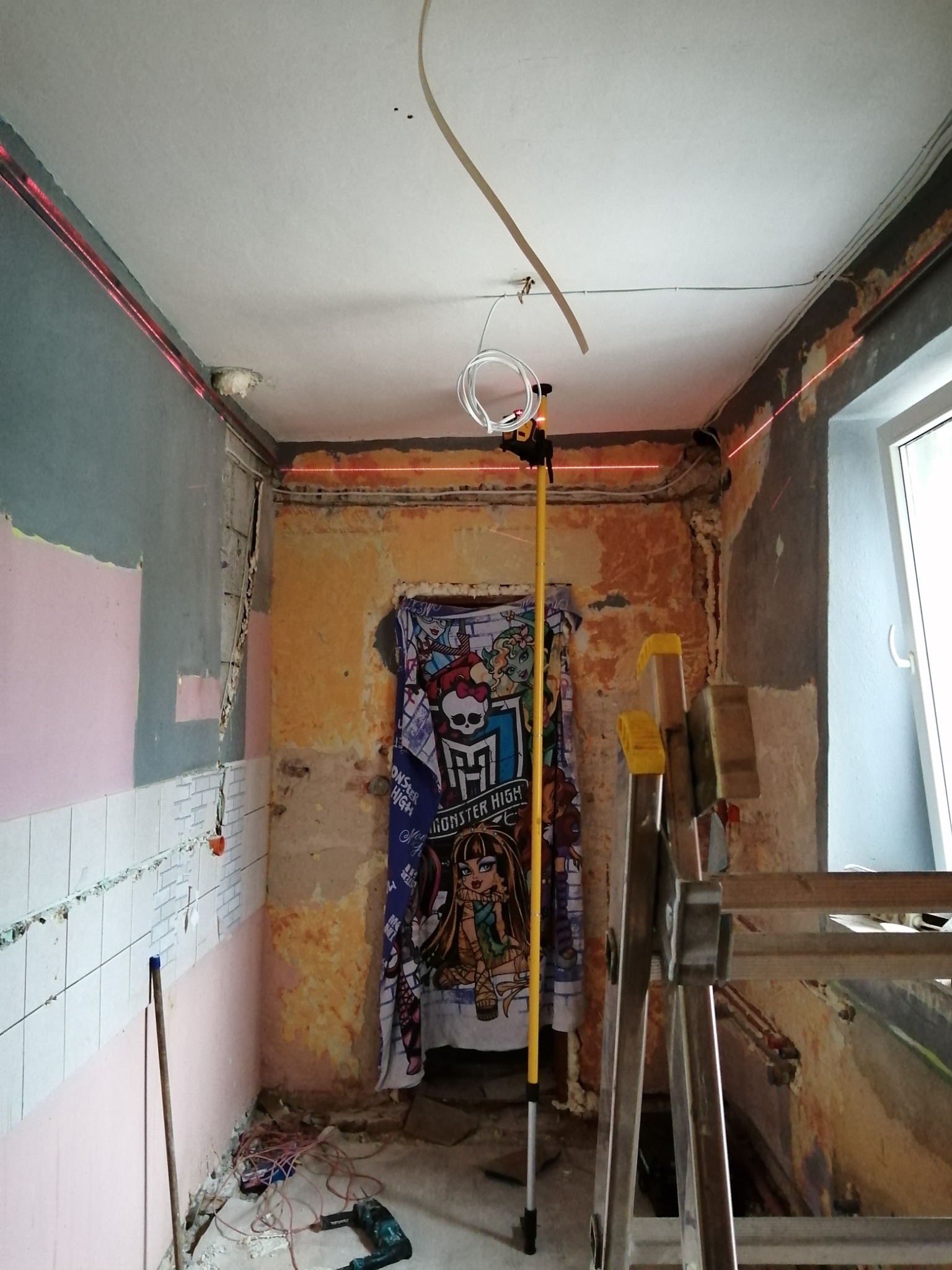 Pokój w trakcie remontu z odsłoniętymi fragmentami ścian, widoczne ślady po starych płytkach, laser krzyżowy wyznacza poziom na ścianach i suficie, drabina aluminiowa i wiszący ręcznik z motywem...