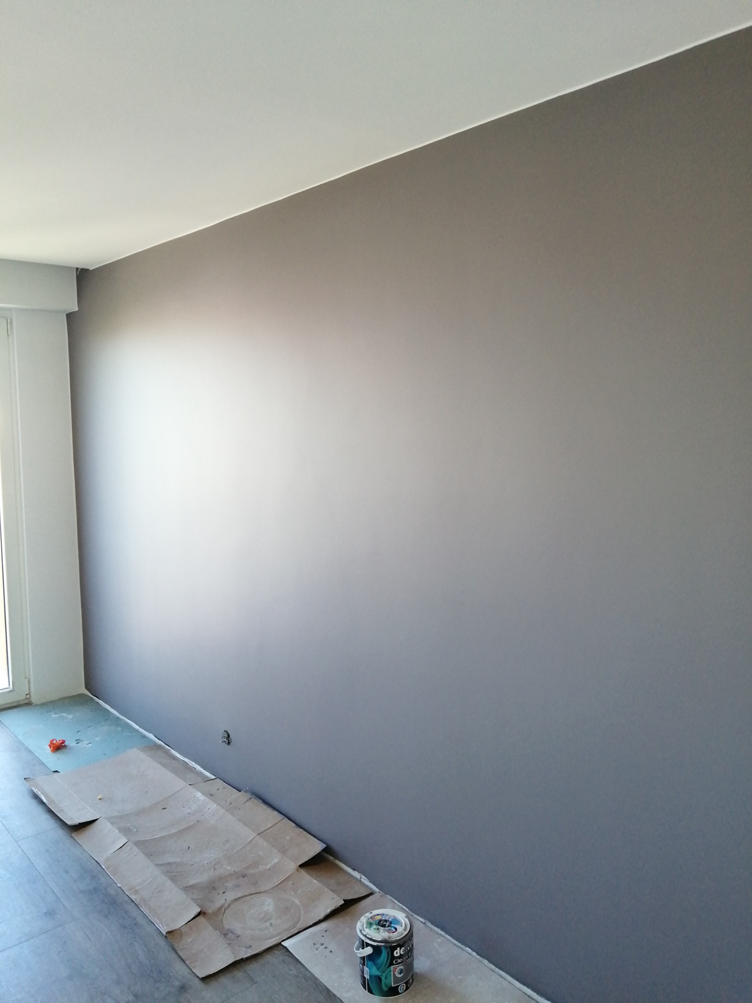 Świeżo pomalowana ściana na szaro w pokoju, zabezpieczona podłoga kartonem, widoczna puszka farby przy ścianie.