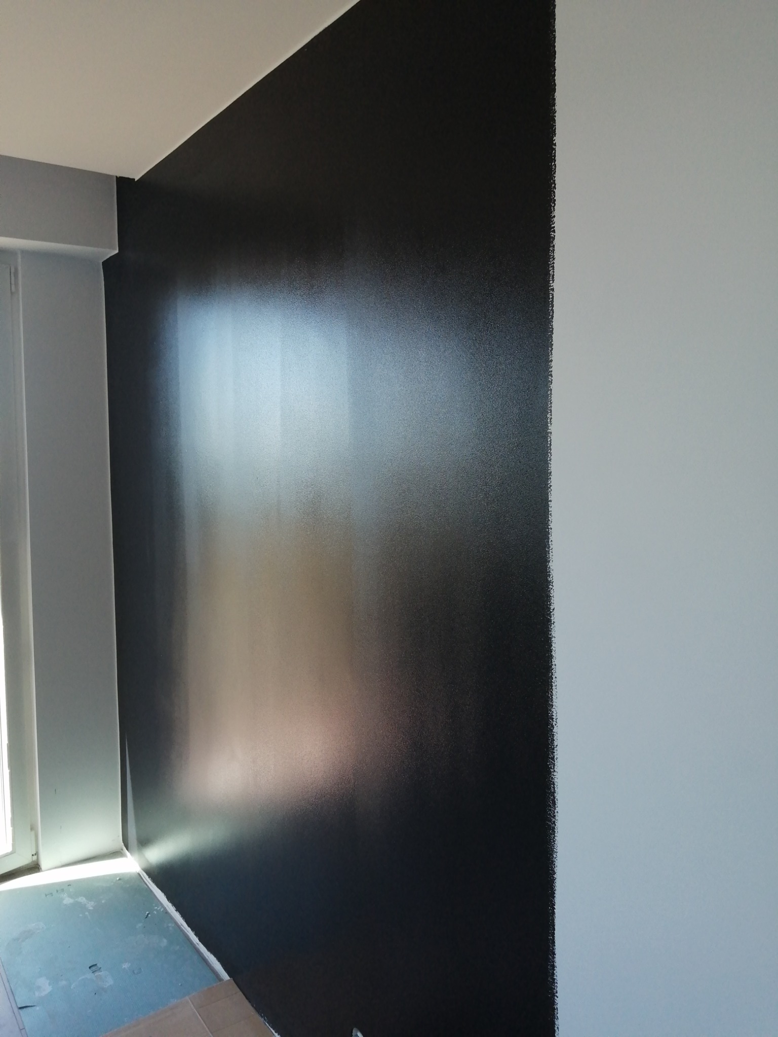 Świeżo pomalowana ściana w pokoju, połowa w kolorze grafitowym, druga połowa biała, widoczne odcięcie kolorów, podłoga zabezpieczona folią malarską, widoczne światło dzienne z okna.