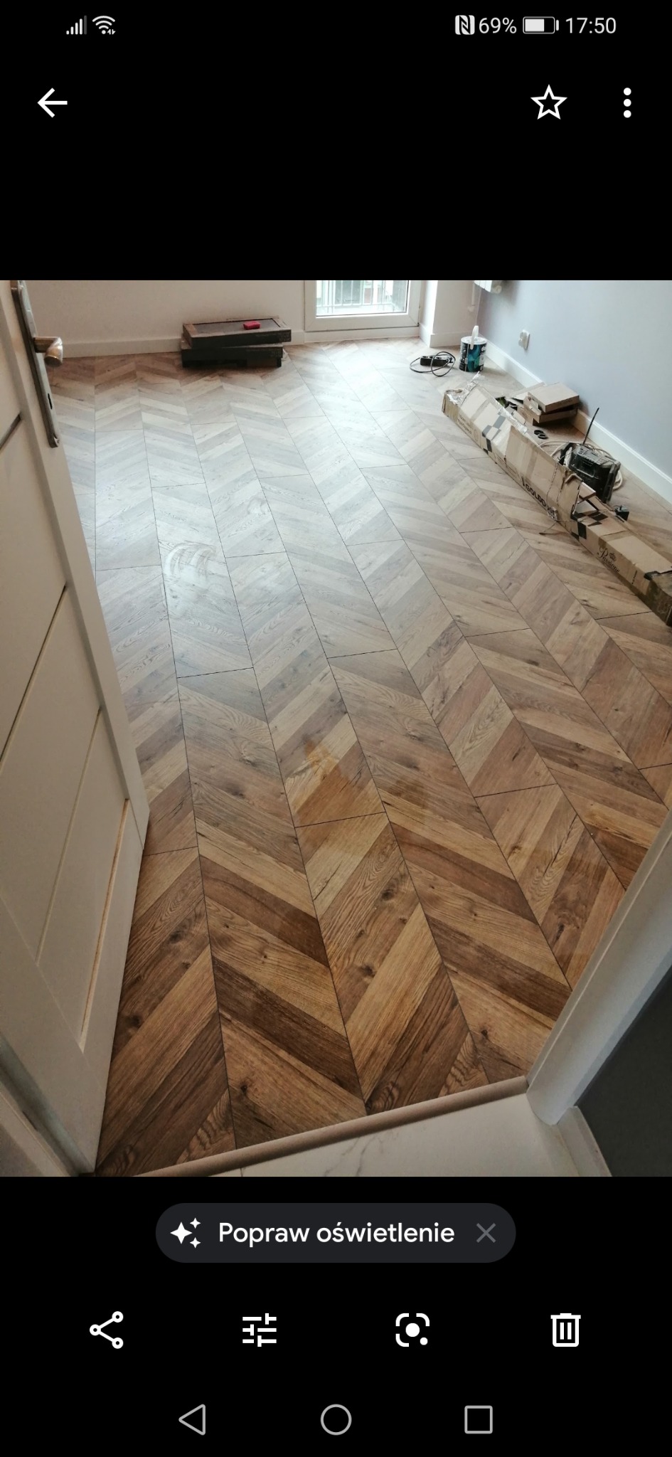 Ułożone panele podłogowe w jodełkę, widoczne opakowania po panelach, fragment drzwi i okna.