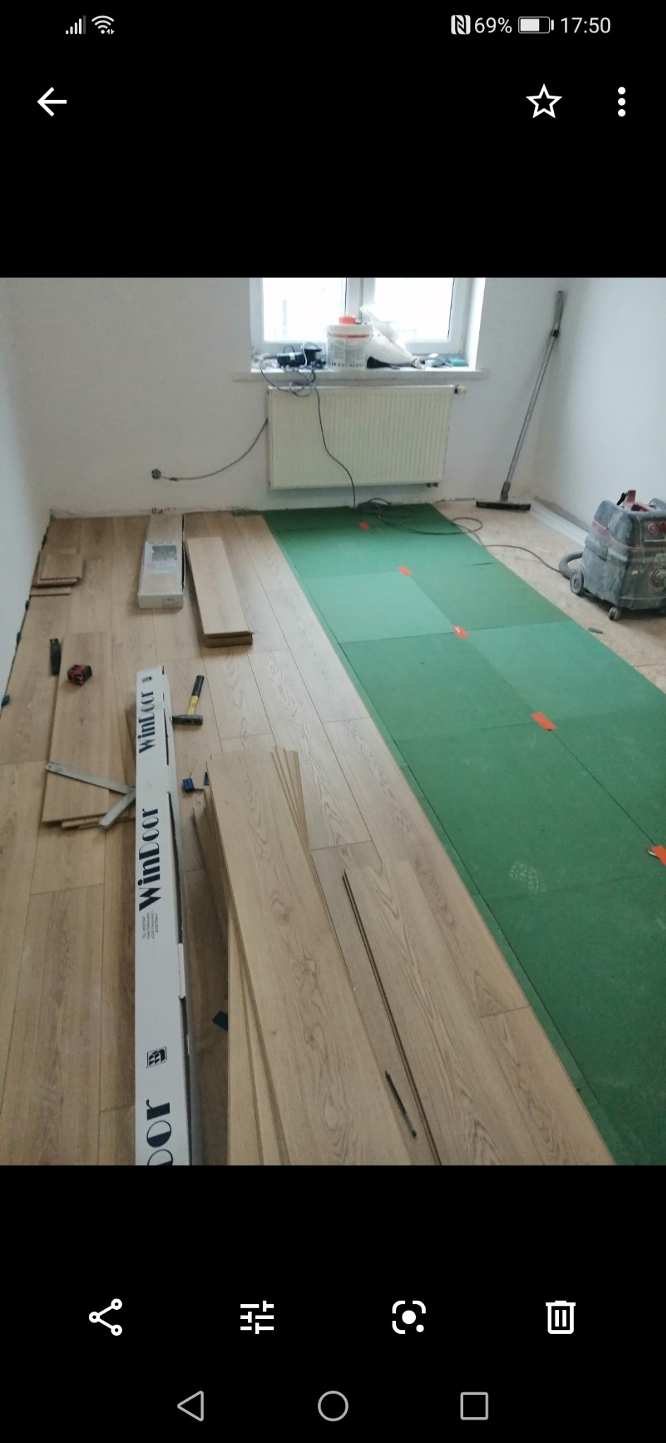 Proces montażu jasnych paneli podłogowych w pokoju, widoczne narzędzia, podkład zielony z pomarańczowymi znacznikami, odkurzacz przemysłowy w tle.