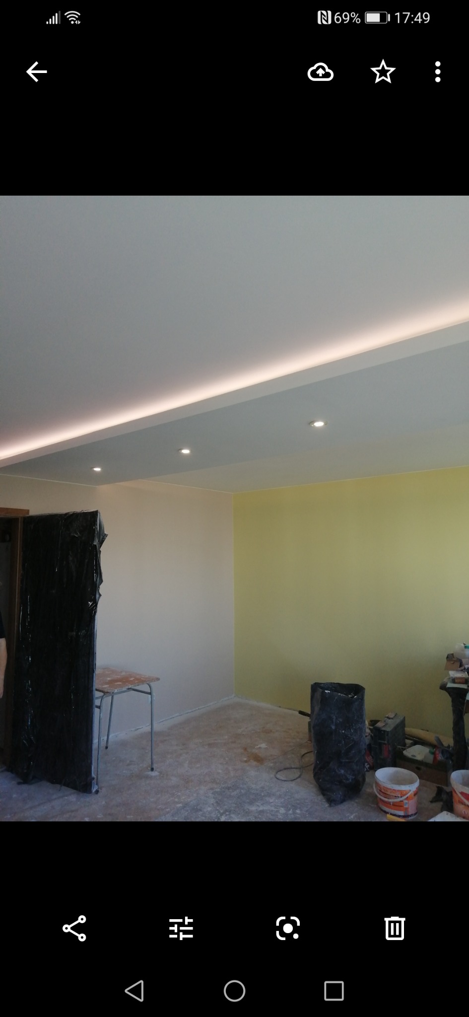 Wnętrze pokoju w trakcie prac wykończeniowych, widoczne pomalowane ściany w dwóch odcieniach, zakryte folią meble, sufit z oświetleniem LED, oraz narzędzia malarskie.