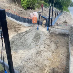 Przygotowanie terenu pod budowę ogrodzenia: widoczny wykop, stabilizacja gruntu czarnymi panelami, wbite słupki ogrodzeniowe, mieszalnik do betonu i pryzma piasku.