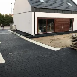 Czarno-szara kostka brukowa ułożona przed nowoczesnym domem z białymi ścianami i drewnianą elewacją, widoczne palety i węże ogrodowe na piasku.