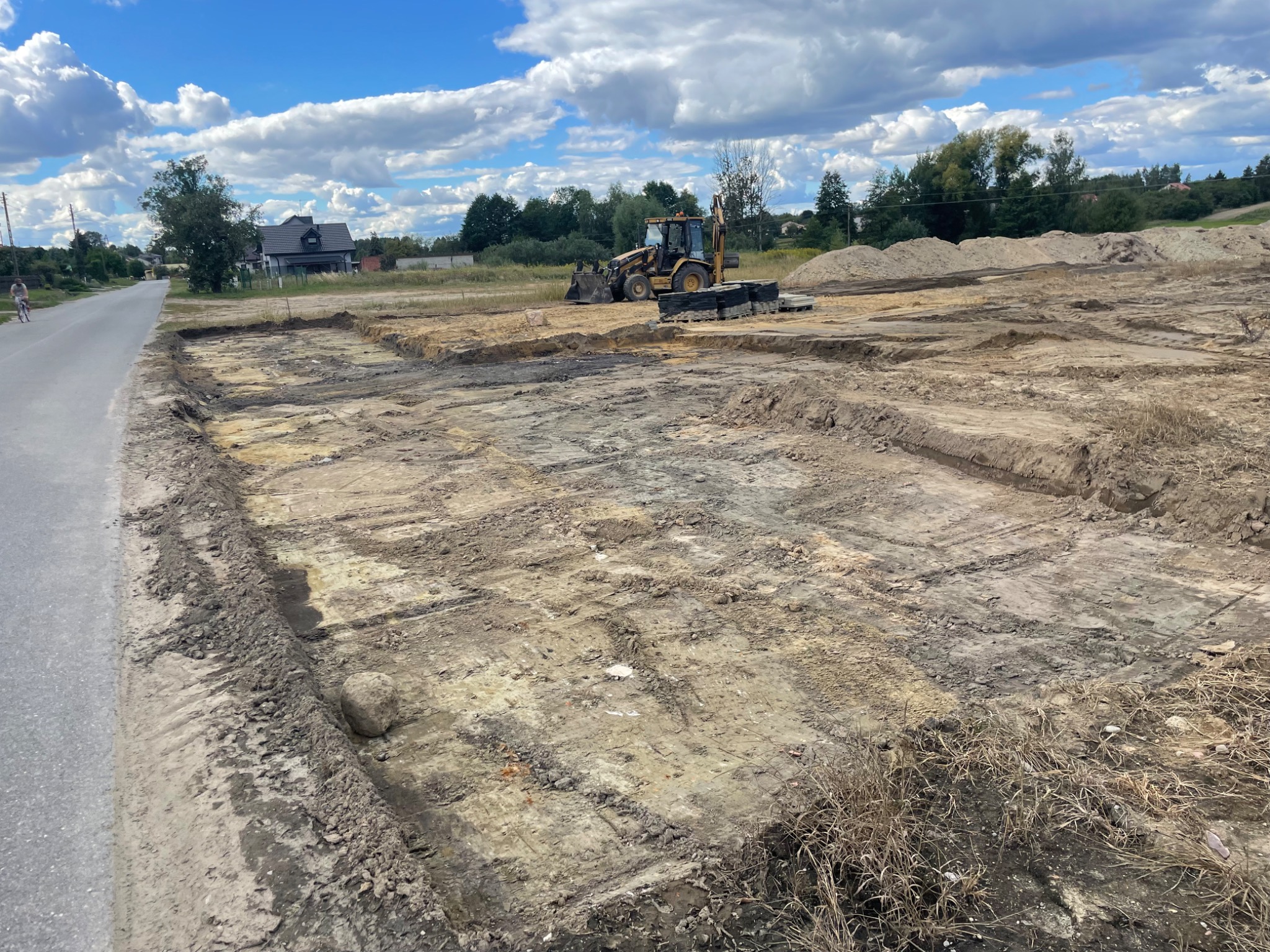 Przygotowany teren pod budowę z widocznymi wykopami, w tle koparko-ładowarka i hałdy ziemi, błękitne niebo z obłokami.