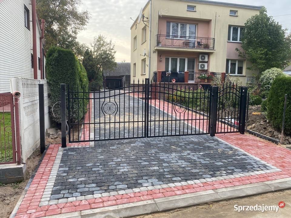 Ułożona kostka brukowa w odcieniach szarości i czerwieni prowadząca do czarnej, ozdobnej bramy wjazdowej przed dwupiętrowym budynkiem z balkonem.