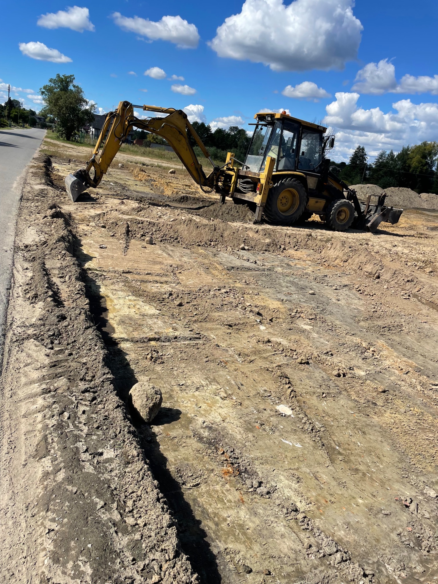 Żółta koparko-ładowarka na placu budowy, widoczne świeżo wykopane rowy w ziemi, błękitne niebo z chmurami w tle, obok asfaltowa droga.