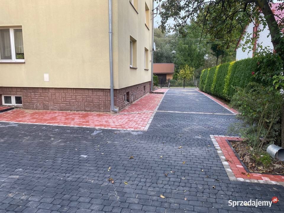 Ułożona z szarej kostki brukowej nawierzchnia z czerwonym obramowaniem, przylegająca do budynku z beżową elewacją i brązową podmurówką, widok na podwórko z zielenią.
