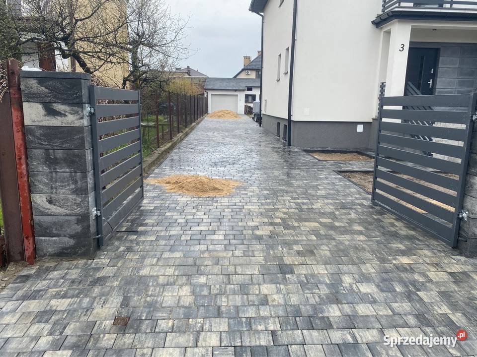 Ułożona szara kostka brukowa na podjeździe z otwartą, metalową bramą w nowoczesnym stylu, widoczne ślady piasku zasypującego szczeliny, dom z numerem 3 w tle.