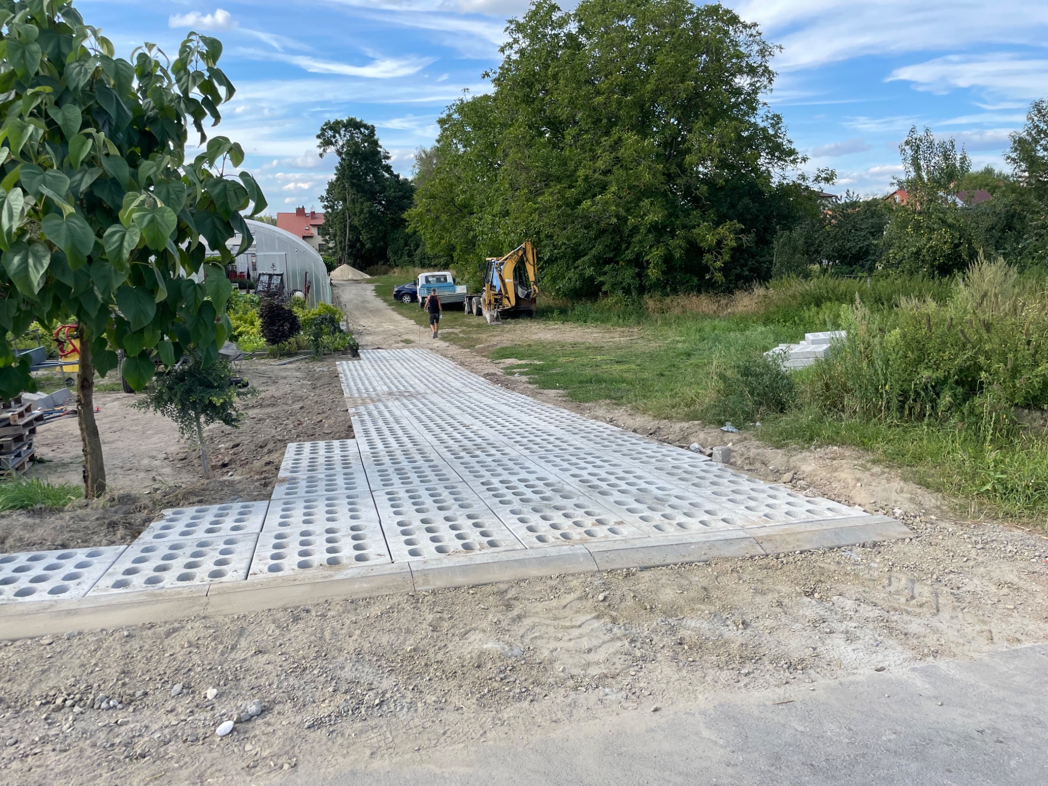 Układanie ażurowych płyt betonowych na podjeździe; widoczny mini-koparka i samochód dostawczy w tle, częściowo ułożona nawierzchnia z kostki ażurowej.