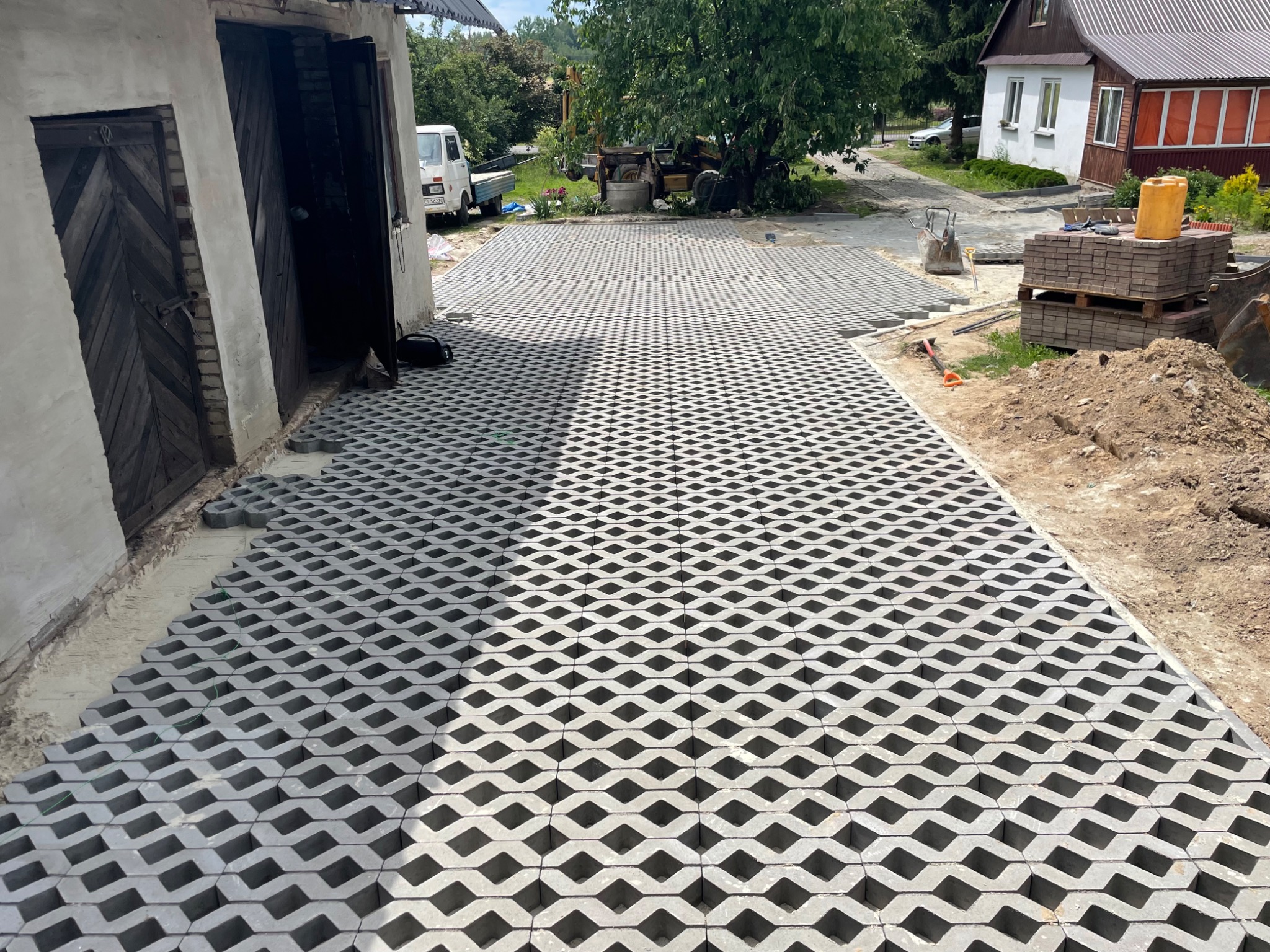 Ułożona częściowo ażurowa nawierzchnia z betonowych płyt w kształcie rombów, z widocznymi stertami kostki i narzędziami w tle, obok budynku gospodarczego i domu jednorodzinnego.