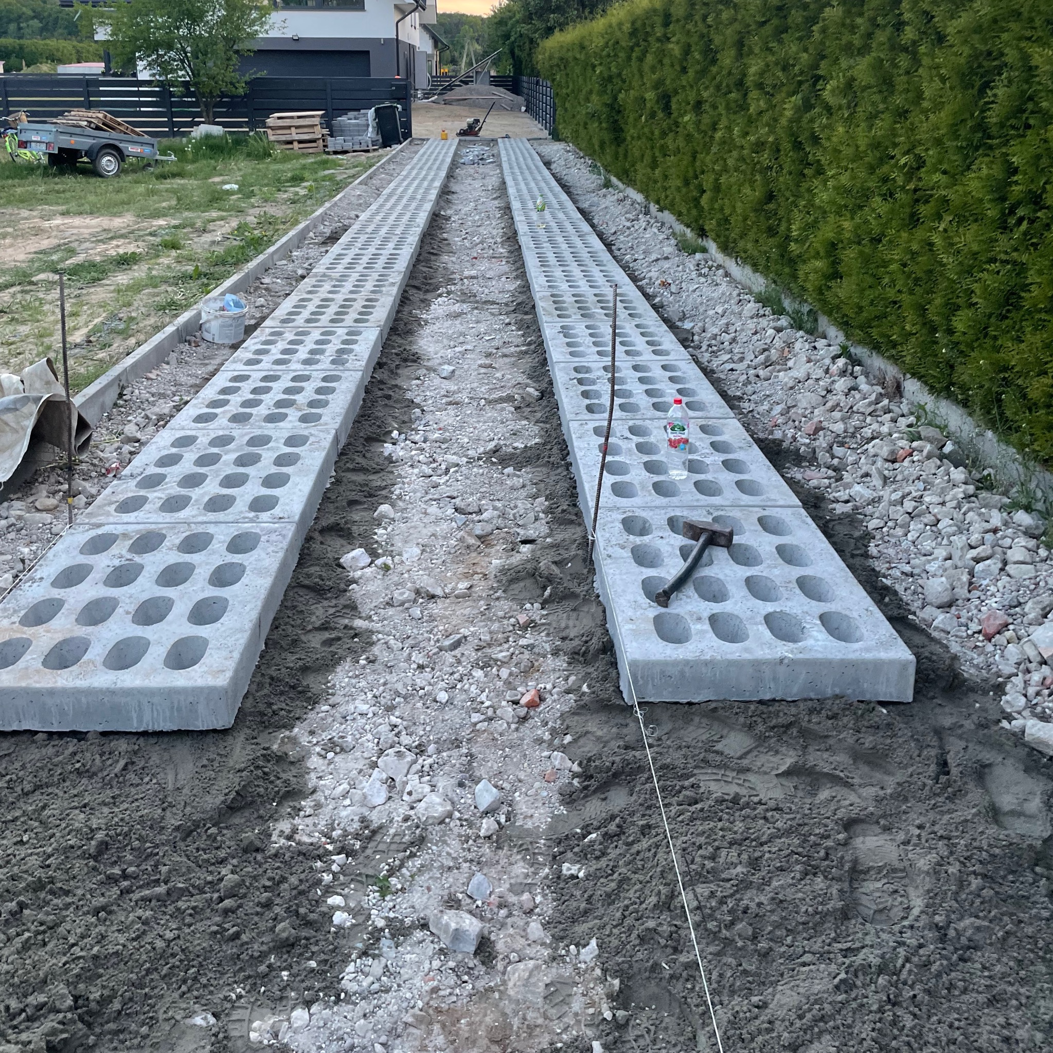 Dwa rzędy betonowych płyt ażurowych ułożone na przygotowanym podłożu z piasku i kamienia, widoczne narzędzia pracy: młotek i sznurek, w tle zielony żywopłot i budynek mieszkalny.
