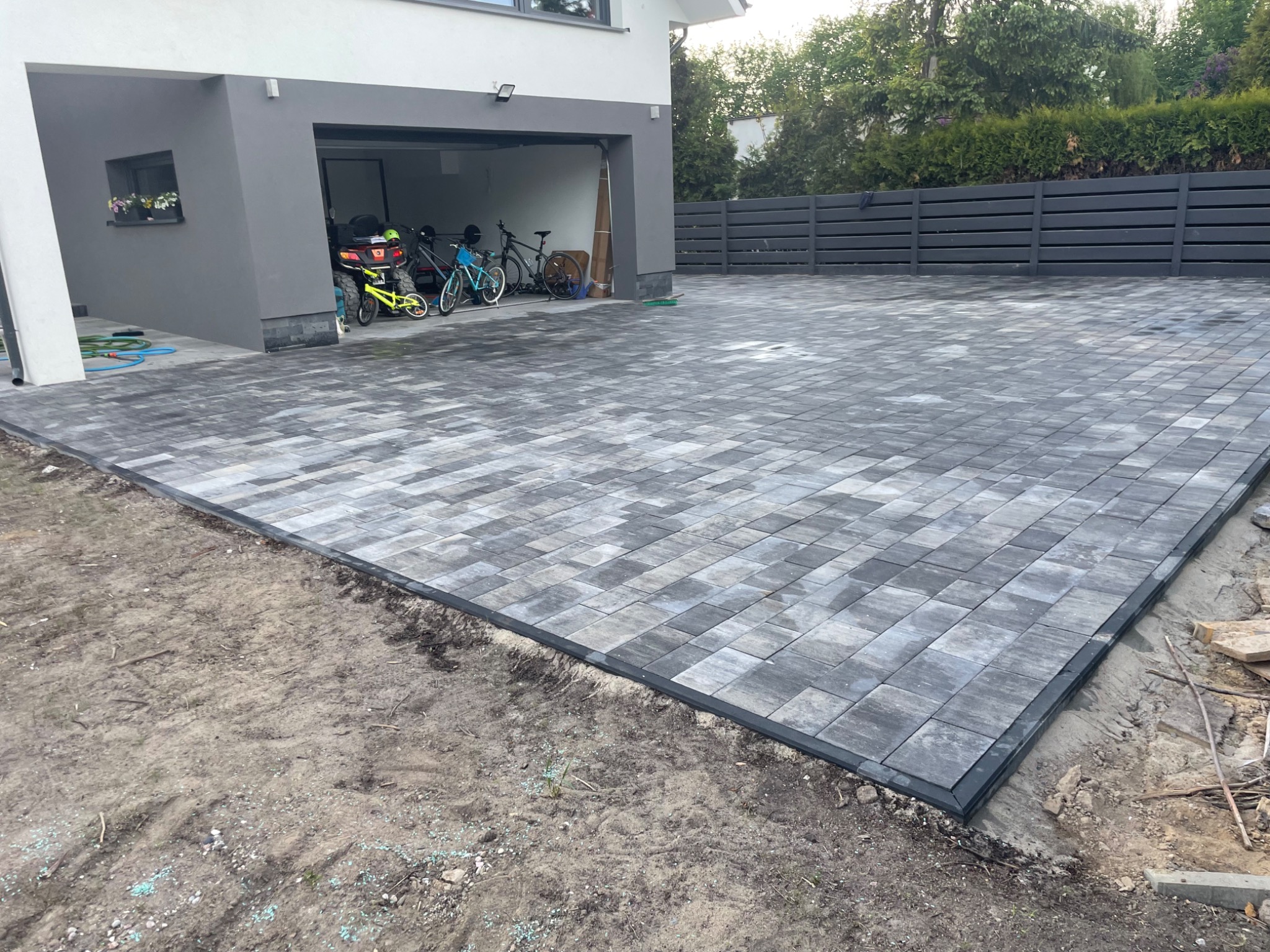 Świeżo ułożona, szara kostka brukowa przed nowoczesnym domem z garażem, obrzeża wykończone, widoczny fragment niezabudowanego terenu z rozsypanymi niebieskimi granulkami na ziemi.
