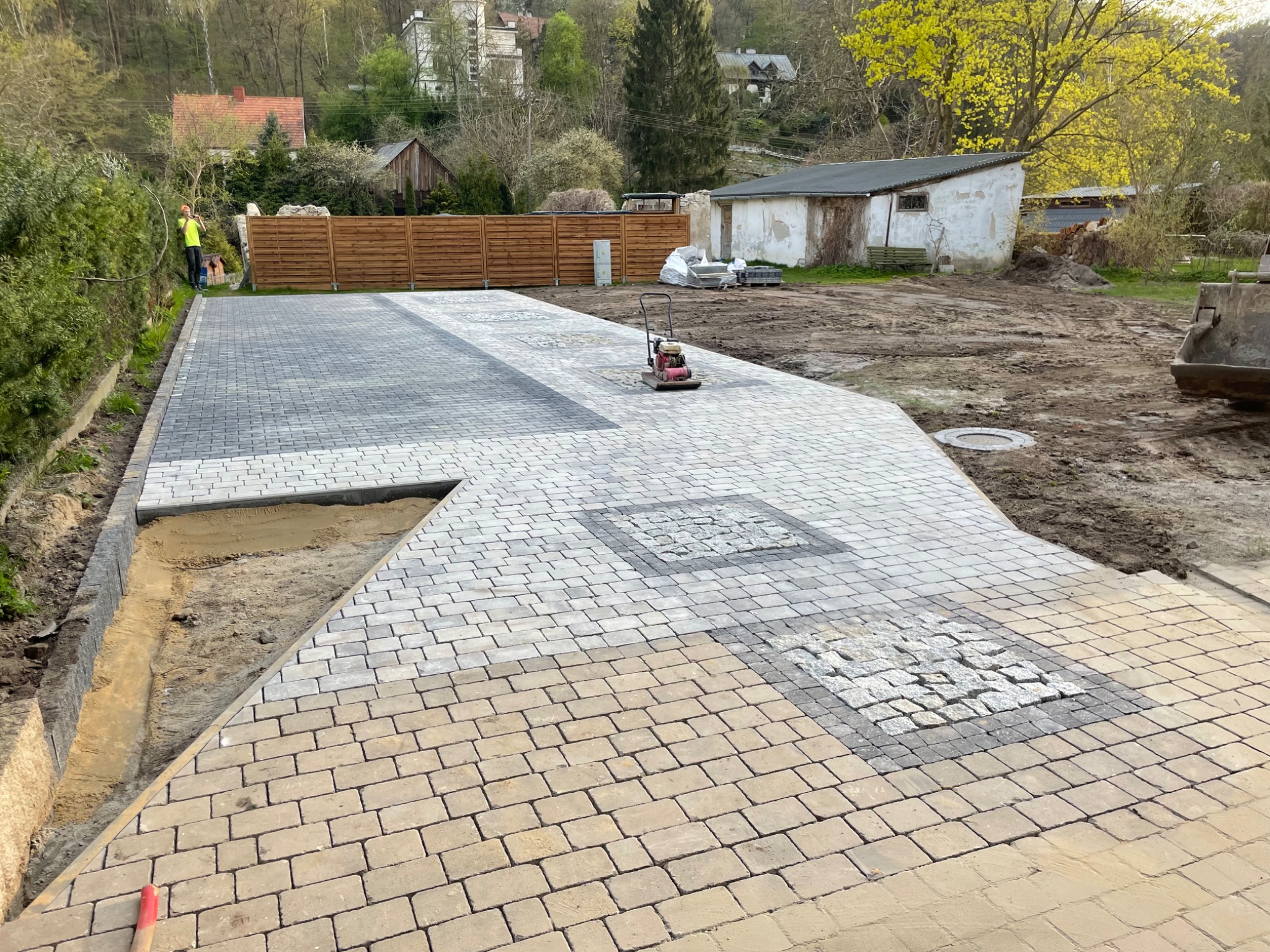 Częściowo ułożona nawierzchnia z kostki brukowej w odcieniach szarości i beżu, z wzorem kwadratów z drobnej kostki, zagęszczarka wibracyjna na powierzchni, widoczny wykop pod obrzeża i pracownik...