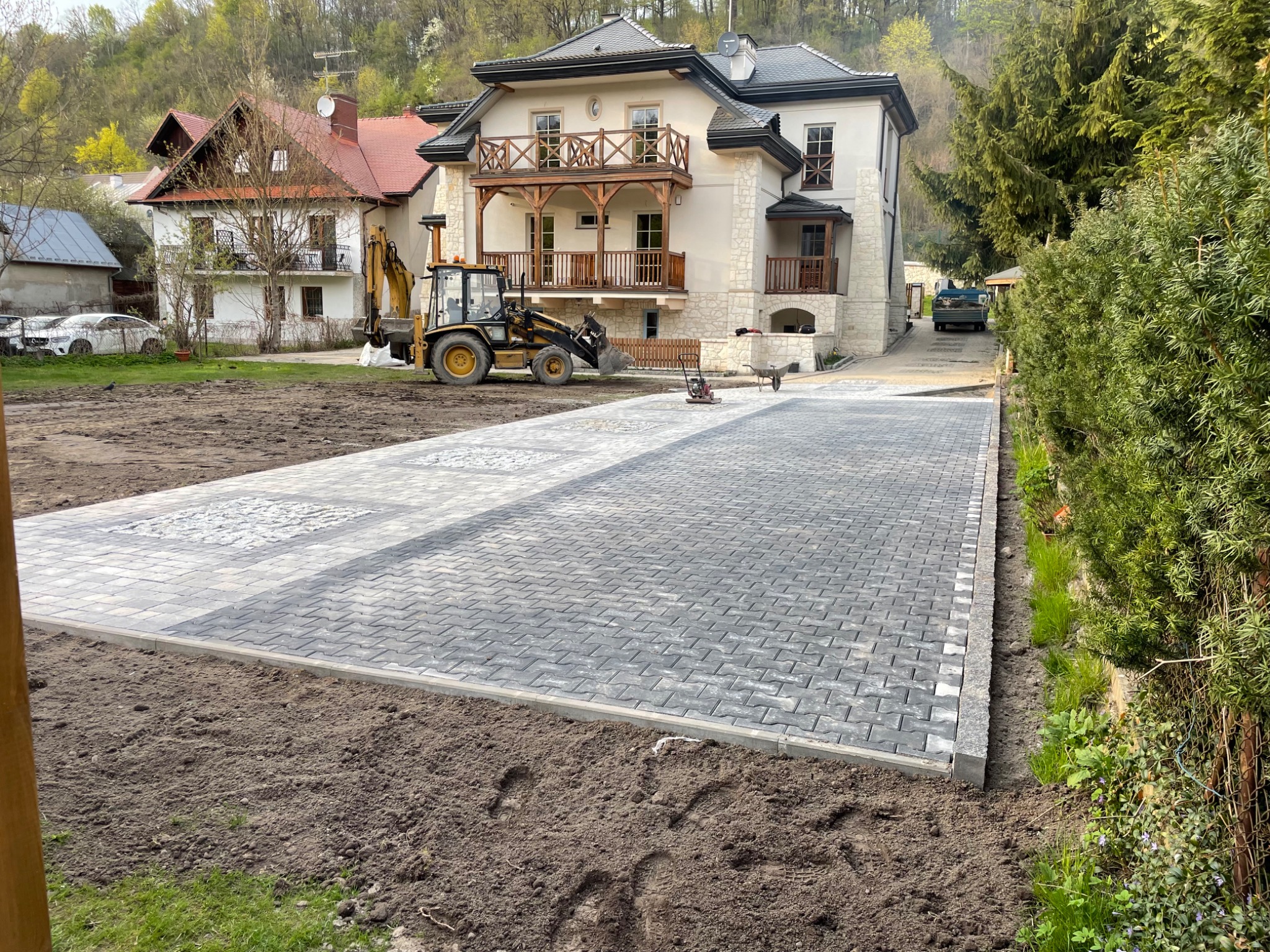 Nowa, szara kostka brukowa ułożona na podjeździe przed domem z beżową elewacją, widoczny traktor z przodu, ubijarka i taczka w tle oraz świeżo nasypana ziemia po bokach