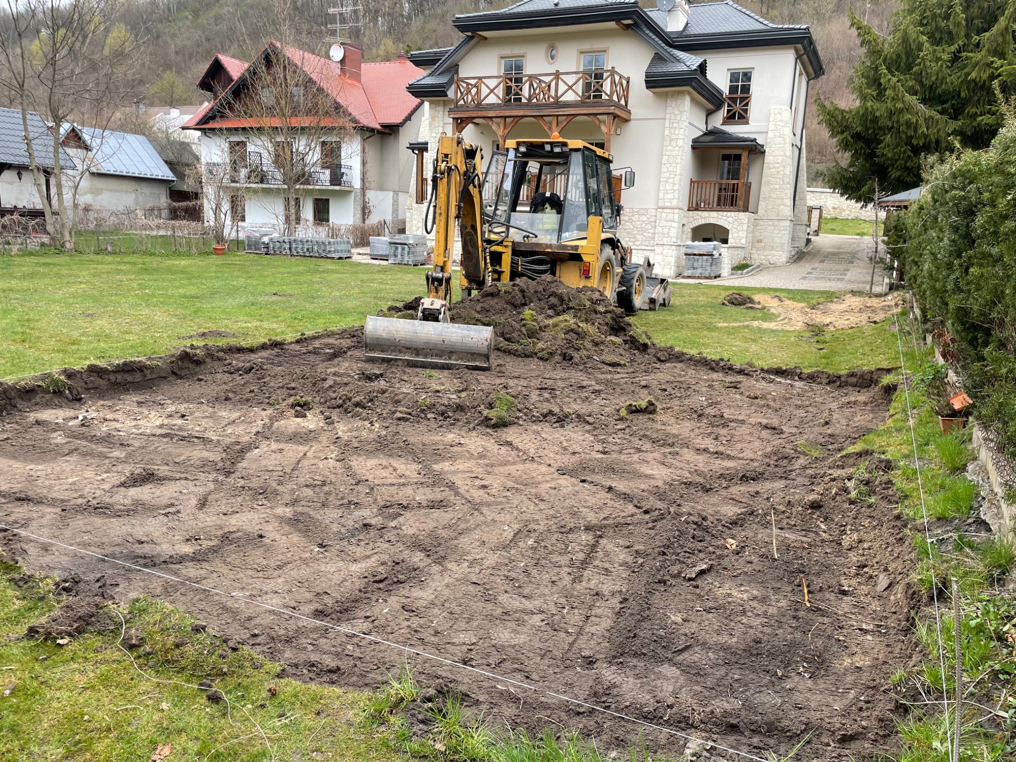 Żółta koparko-ładowarka na działce z przygotowanym terenem pod budowę, widoczne białe sznurki wyznaczające granice, w tle domy jednorodzinne i drzewa.