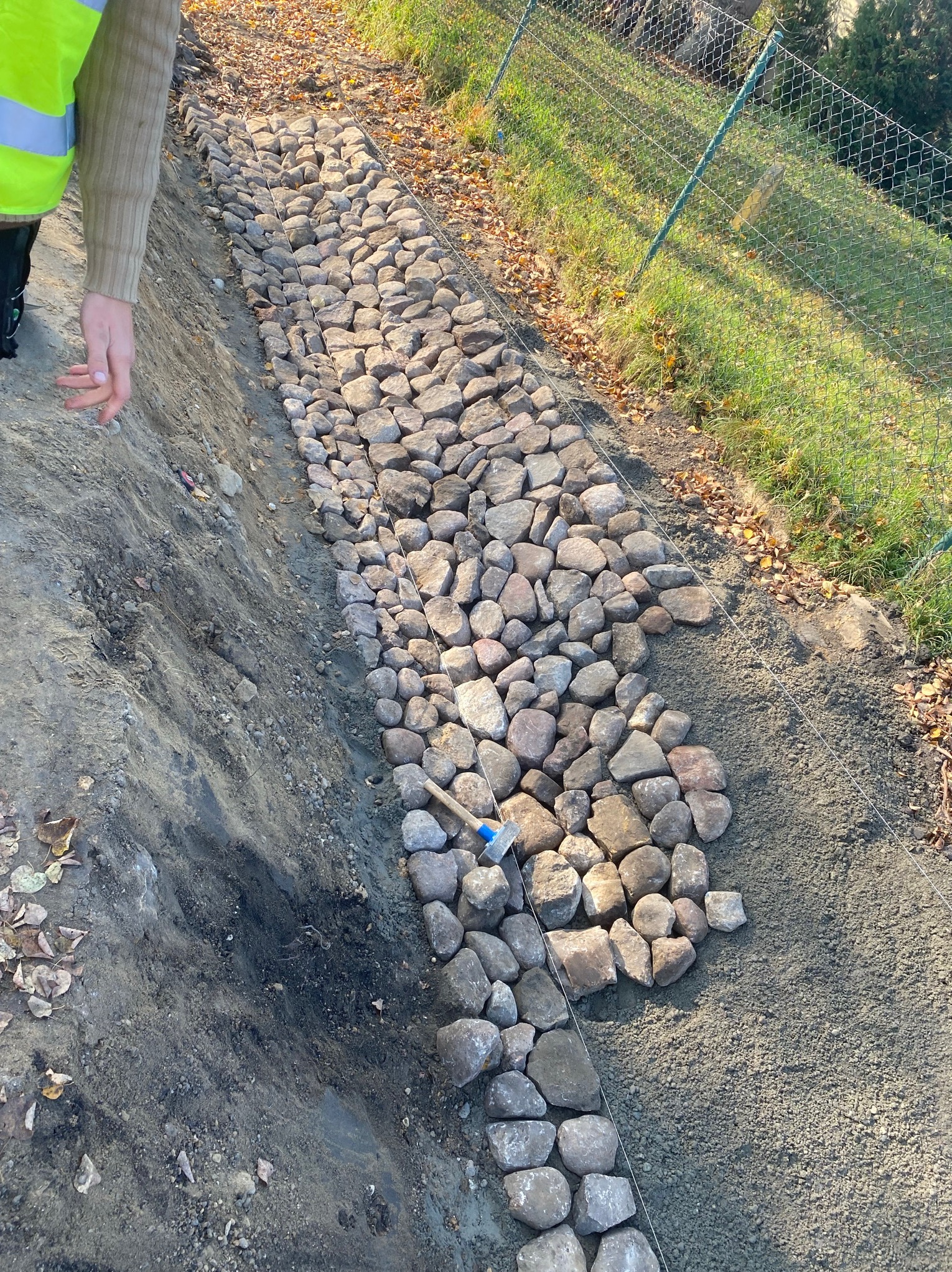 Budowa kamiennego murku oporowego na skarpie; widoczne ułożone kamienie, napięty sznurek wyznaczający linię, oraz młotek brukarski i fragment sylwetki pracownika w odblaskowej kamizelce.
