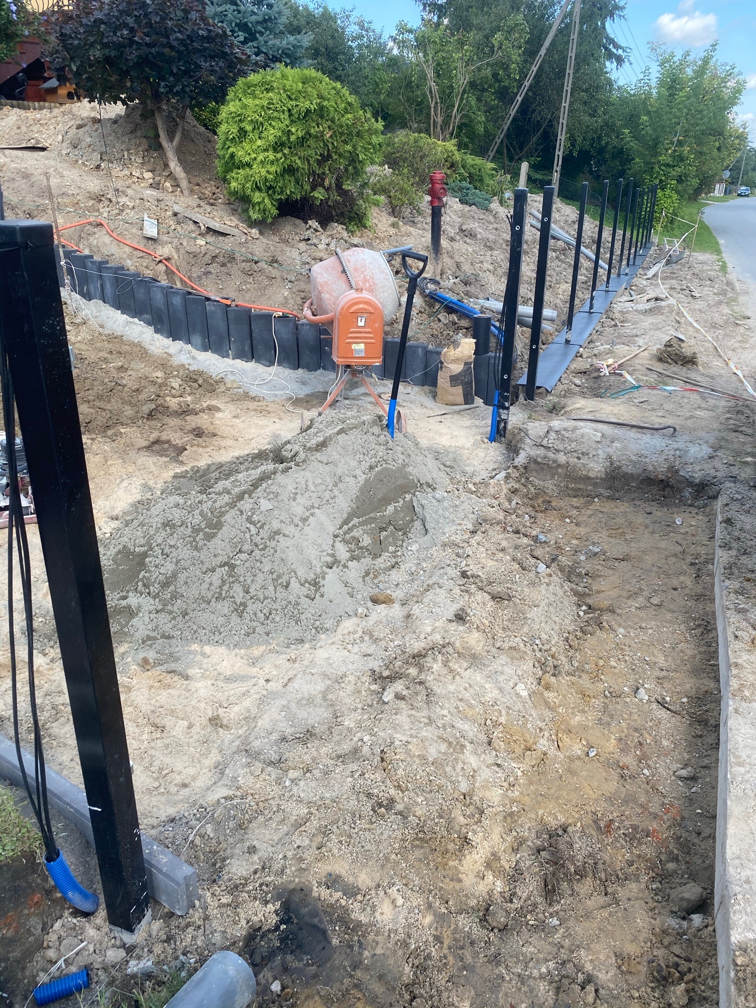 Przygotowanie terenu pod budowę ogrodzenia: widoczny wykop, stabilizacja gruntu czarnymi panelami, wbite słupki ogrodzeniowe, mieszalnik do betonu i pryzma piasku.