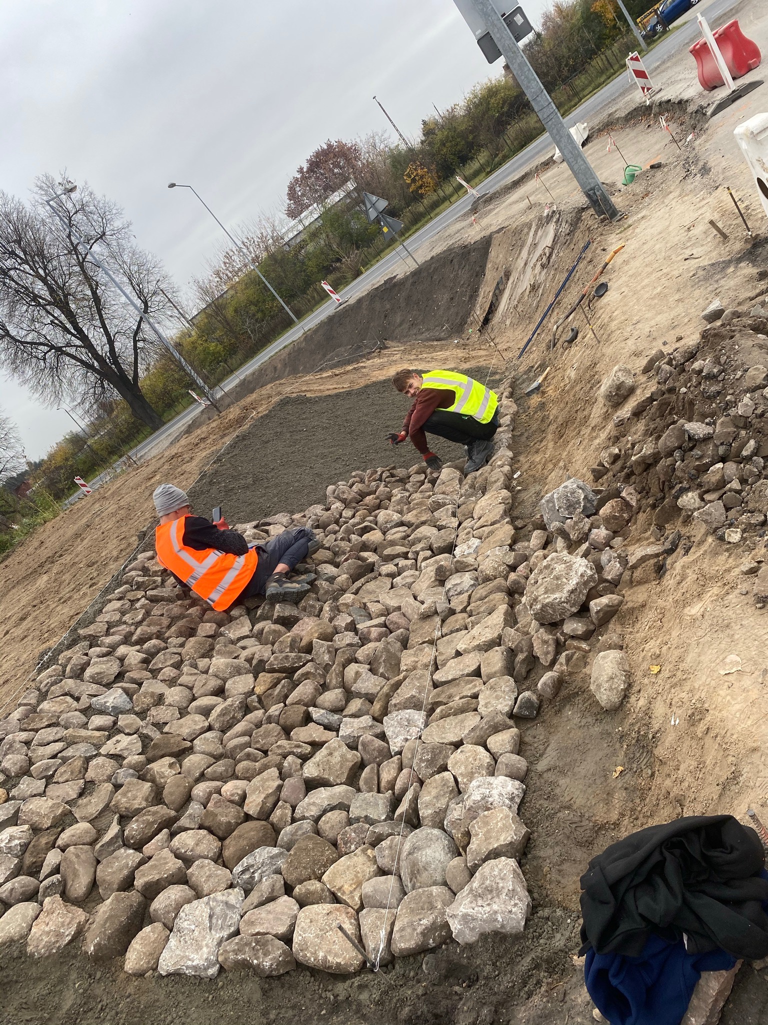 Układanie kamiennej nawierzchni przy drodze przez dwóch pracowników w odblaskowych kamizelkach; jeden siedzi na ułożonych kamieniach z telefonem, drugi klęczy i układa kolejne kamienie; widoczne...
