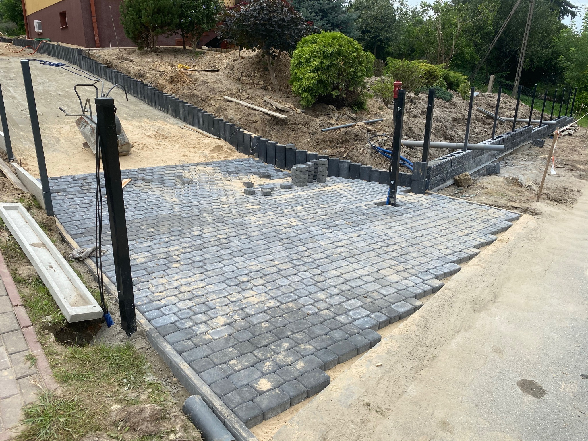 Fragment podjazdu z szarej kostki brukowej w trakcie układania, widoczne przygotowane podłoże, obrzeża i niezabudowane kostki, w tle teren w trakcie niwelacji z palikami ogrodzeniowymi.
