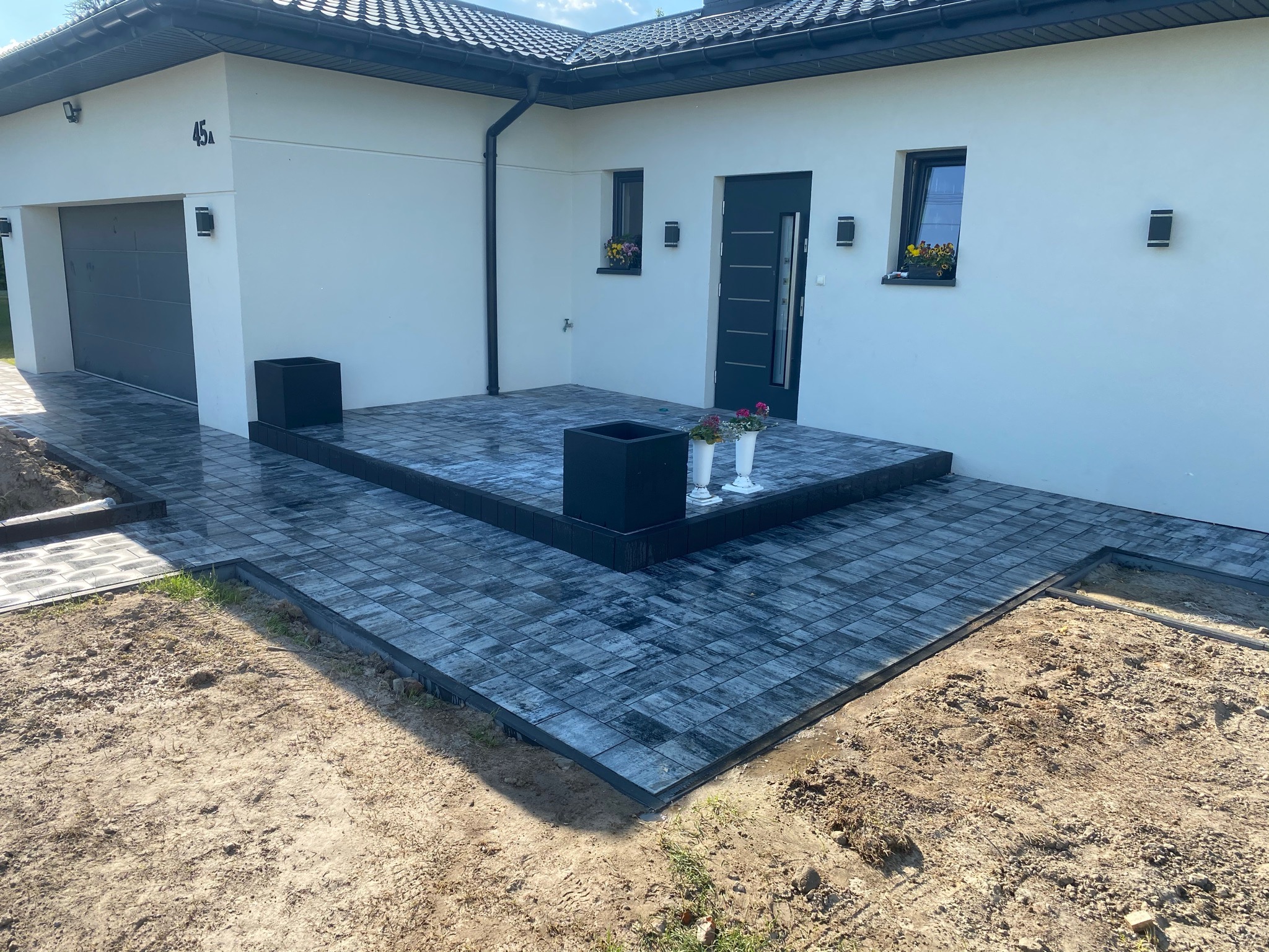 Nowoczesna posesja z szarym brukiem ułożonym przed domem z garażem i czarną stolarką okienno-drzwiową, widoczne donice z kwiatami i fragmenty niezakończonych prac ziemnych.