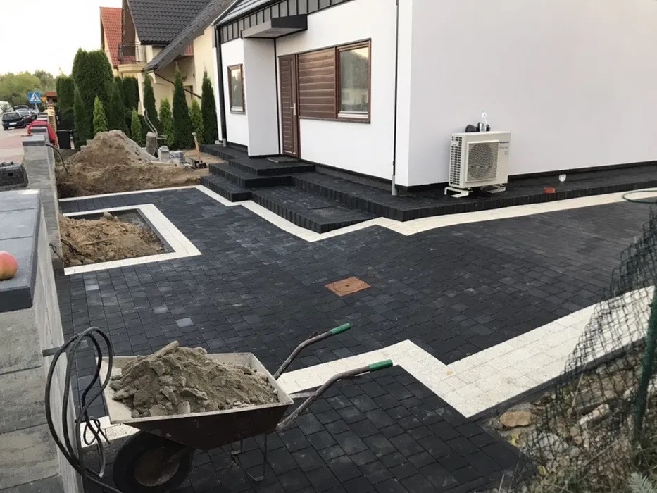 Ułożona czarna kostka brukowa z białymi akcentami przed nowoczesnym domem, taczka z ziemią i wykop w tle, widok na schody wejściowe i klimatyzator.