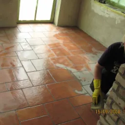 Kobieta w żółtych rękawicach czyści gąbką nowo położoną, brązową terakotę na podłodze pomieszczenia podczas prac wykończeniowych, widoczne resztki fugi, ściany przygotowane do malowania, okno...