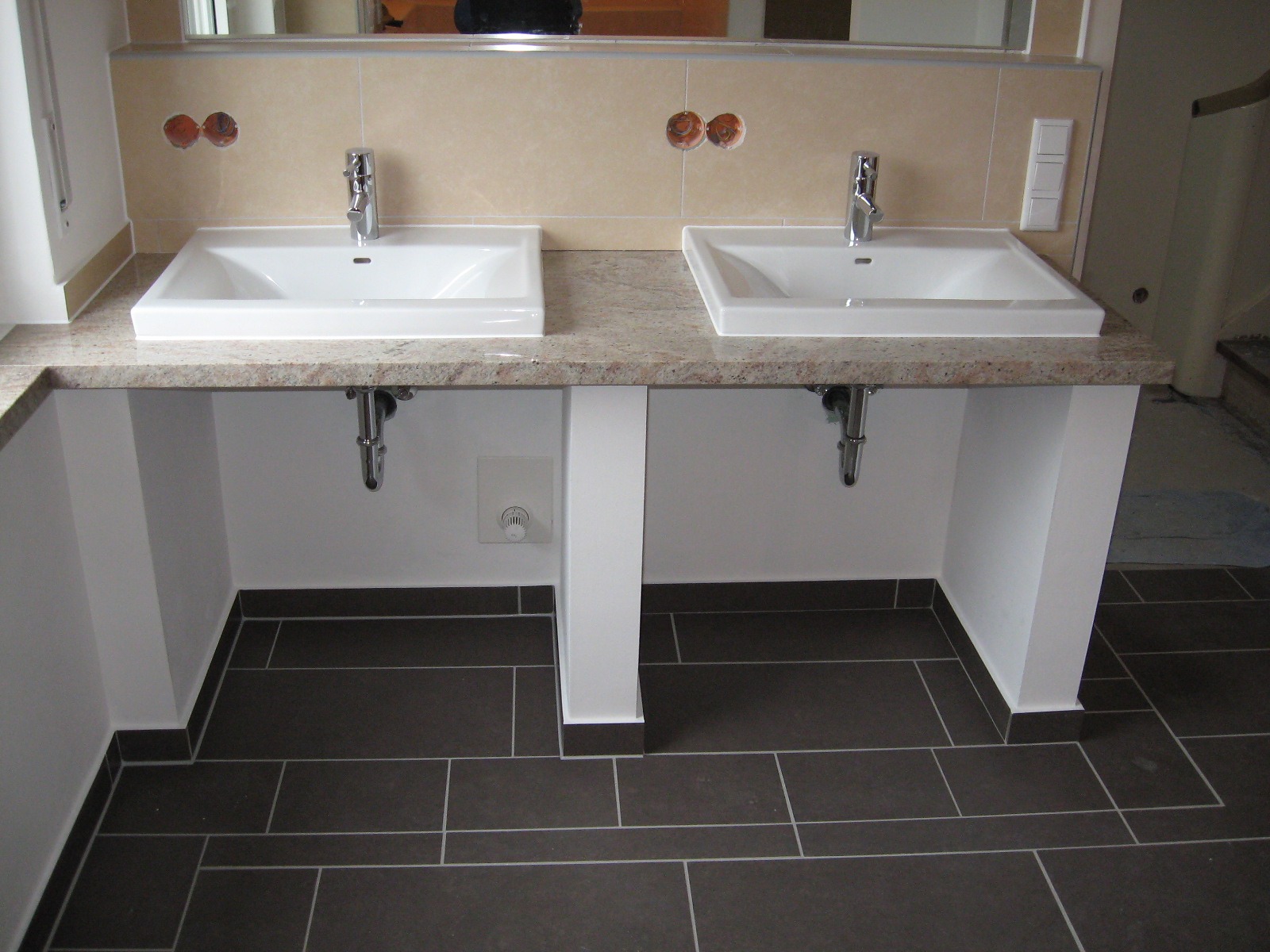 Dwie umywalki ceramiczne o kwadratowym kształcie, zamontowane na blacie z beżowego kamienia z delikatnym wzorem, na tle beżowych płytek ściennych z widocznymi otworami na armaturę, ciemnobrązowa...