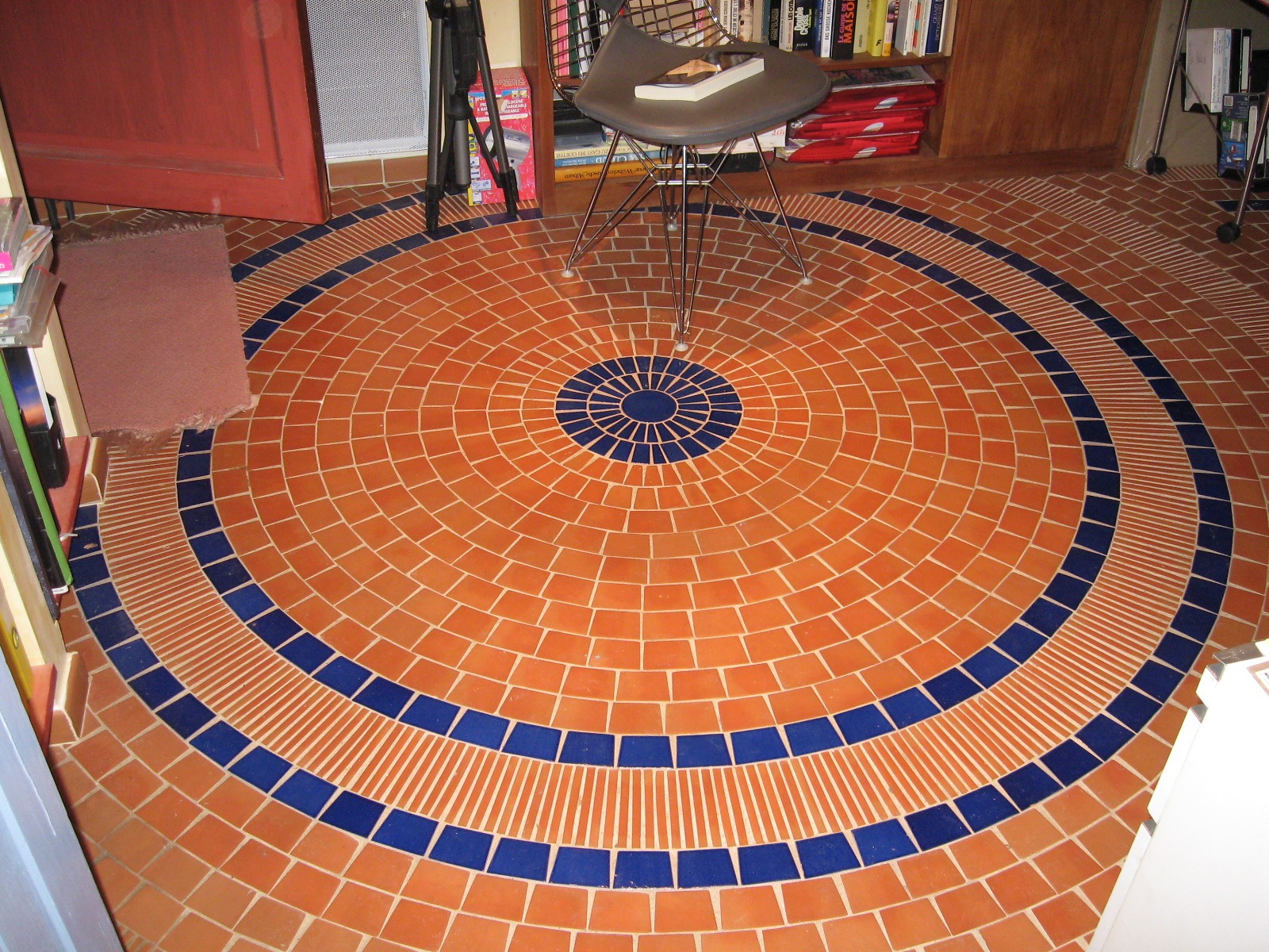Okrągła podłoga z terakoty i niebieskich płytek tworzących koncentryczne wzory, z krzesłem i książką w tle.