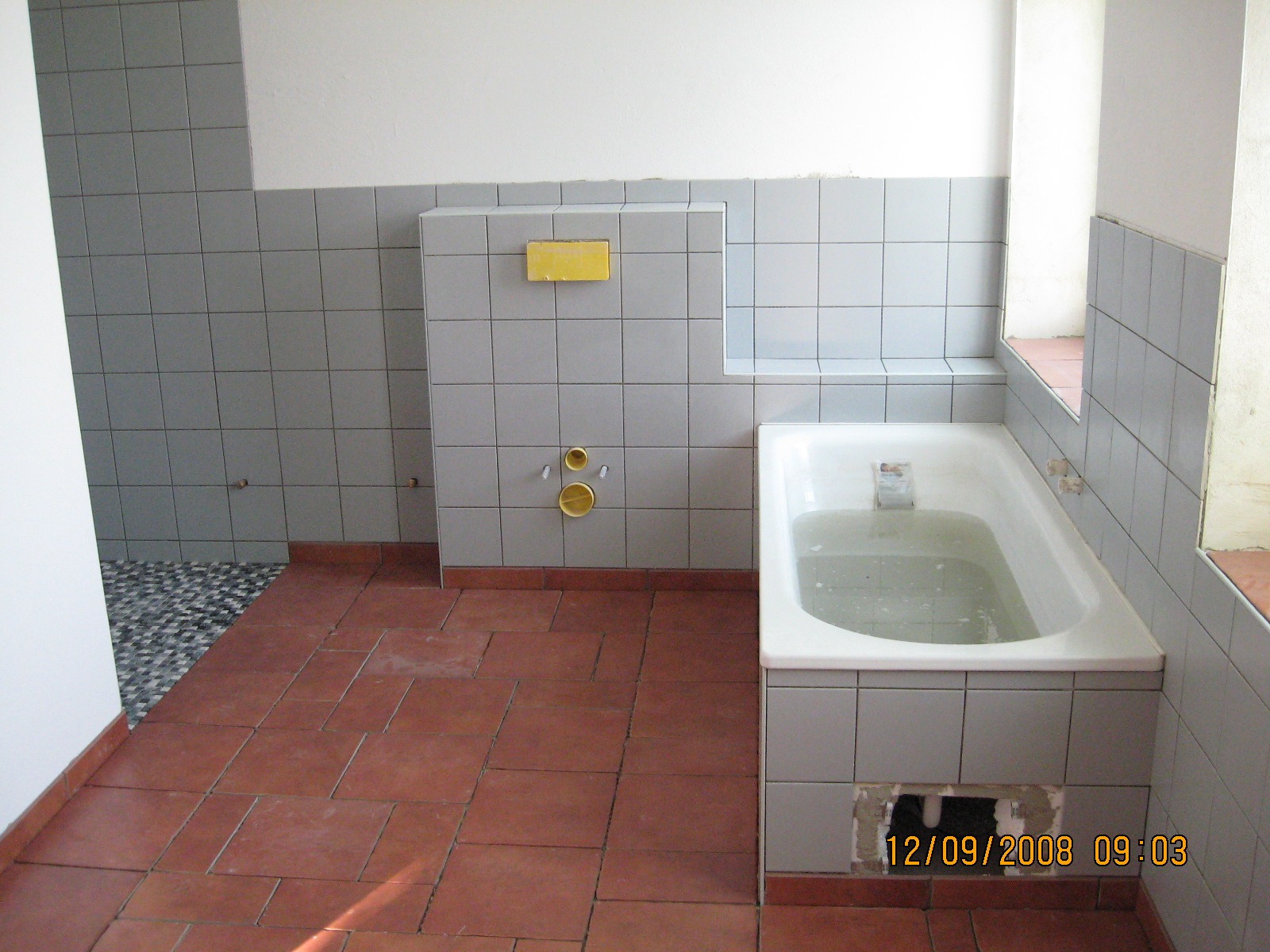 Łazienka w trakcie prac wykończeniowych, widoczne szare płytki na ścianach, terakota na podłodze, wanna wypełniona wodą oraz otwór w obudowie wanny, przygotowane przyłącza wodne i fragment mozaiki...