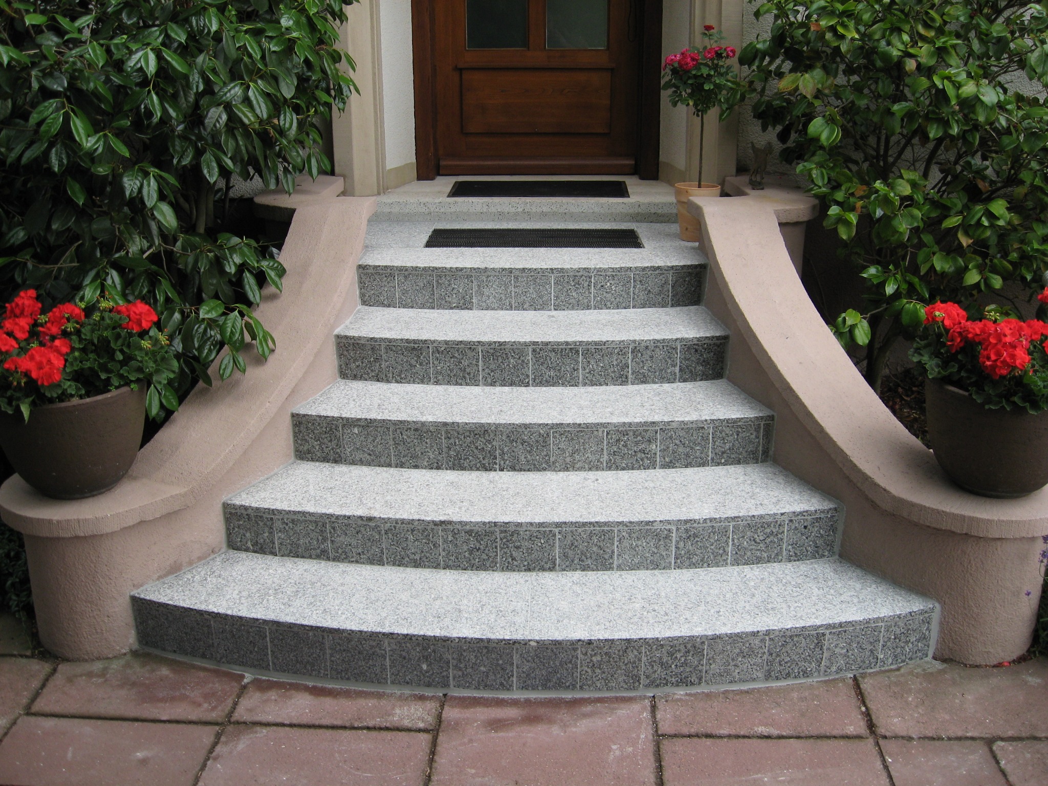Granitowe schody wejściowe z jasnoszarej mozaiki, otoczone niskimi ściankami z donicami z czerwonymi pelargoniami, prowadzące do drewnianych drzwi.