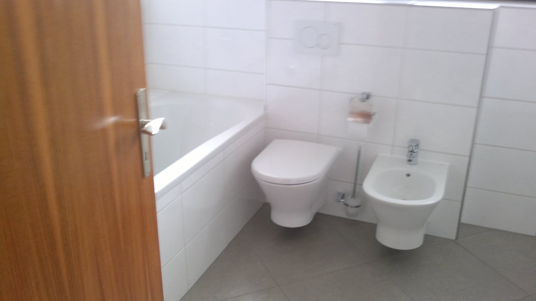 Standardowa, biała łazienka z wanną, toaletą i bidetem, widok zza częściowo otwartych drewnianych drzwi.