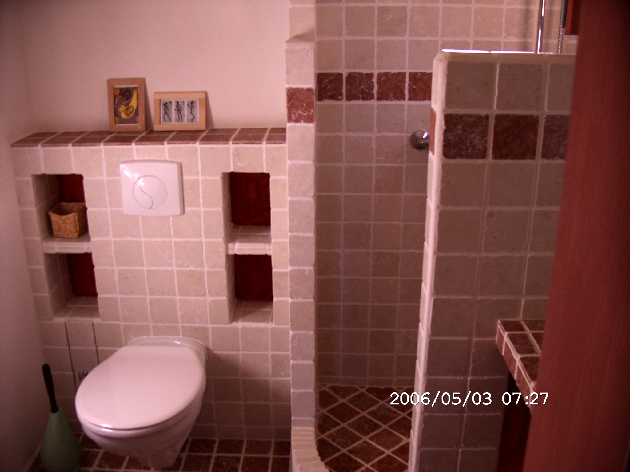 Wykończona łazienka z beżowymi płytkami, wnękami na ścianie, toaletą i kabiną prysznicową z brązowymi akcentami, ozdobiona obrazkami w ramkach i koszykiem.