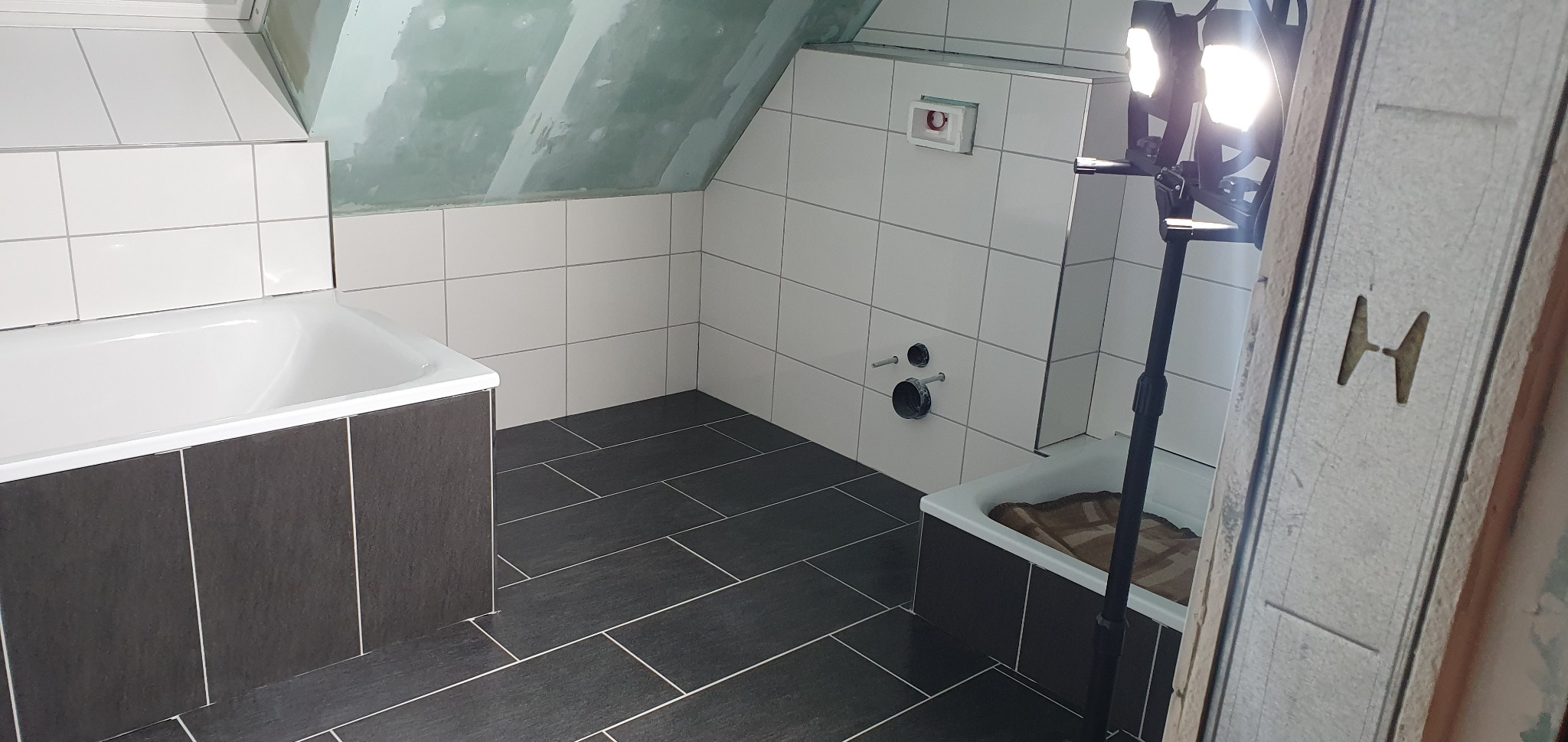 Łazienka w trakcie prac wykończeniowych: białe płytki ścienne, grafitowa podłoga, widoczne rury i instalacja podtynkowa, oświetlenie budowlane i surowy fragment ściany pod skosem.