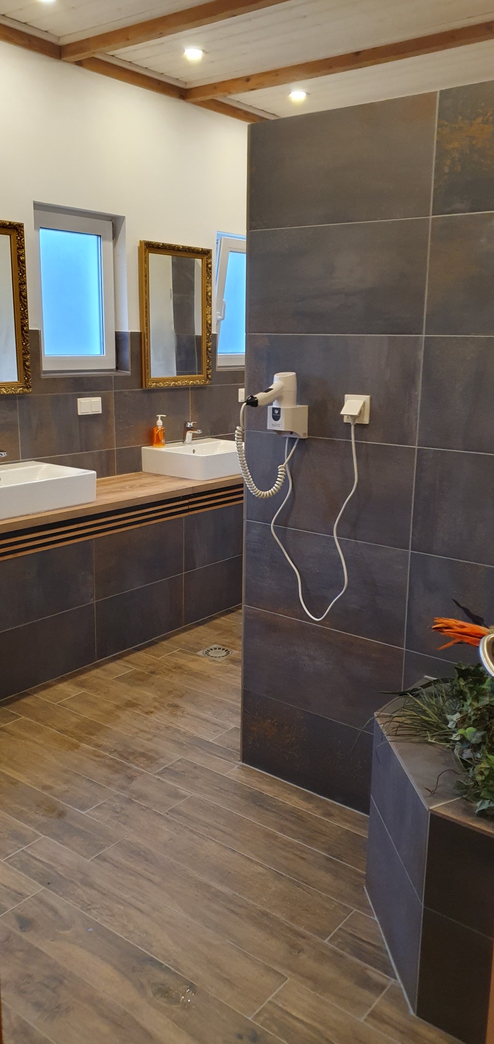 Nowoczesna łazienka z dwoma umywalkami, lustrami w złotych ramach, drewnianą podłogą i ciemnymi płytkami, wyposażona w suszarkę do włosów zamontowaną na ścianie.
