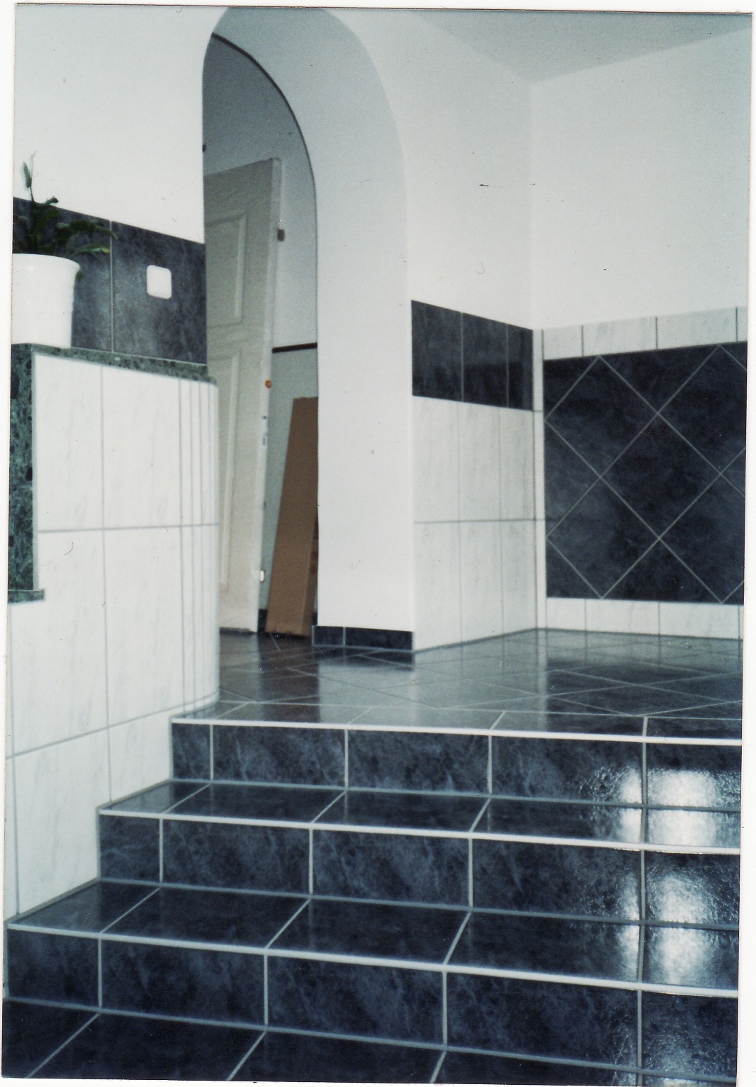 Wykończona łazienka z podwyższonym poziomem podłogi, wyłożona ciemnymi, marmurkowymi płytkami z jasnymi fugami, kontrastująca z białymi ścianami i elementami dekoracyjnymi, w tle widoczne otwarte...